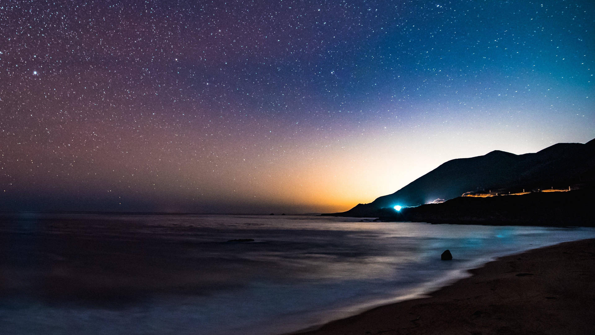 雅虎星空海灘 壁紙 — 繁星點點的夜空在平靜的海洋上閃耀，遠處有山脈輪廓和海岸燈光。