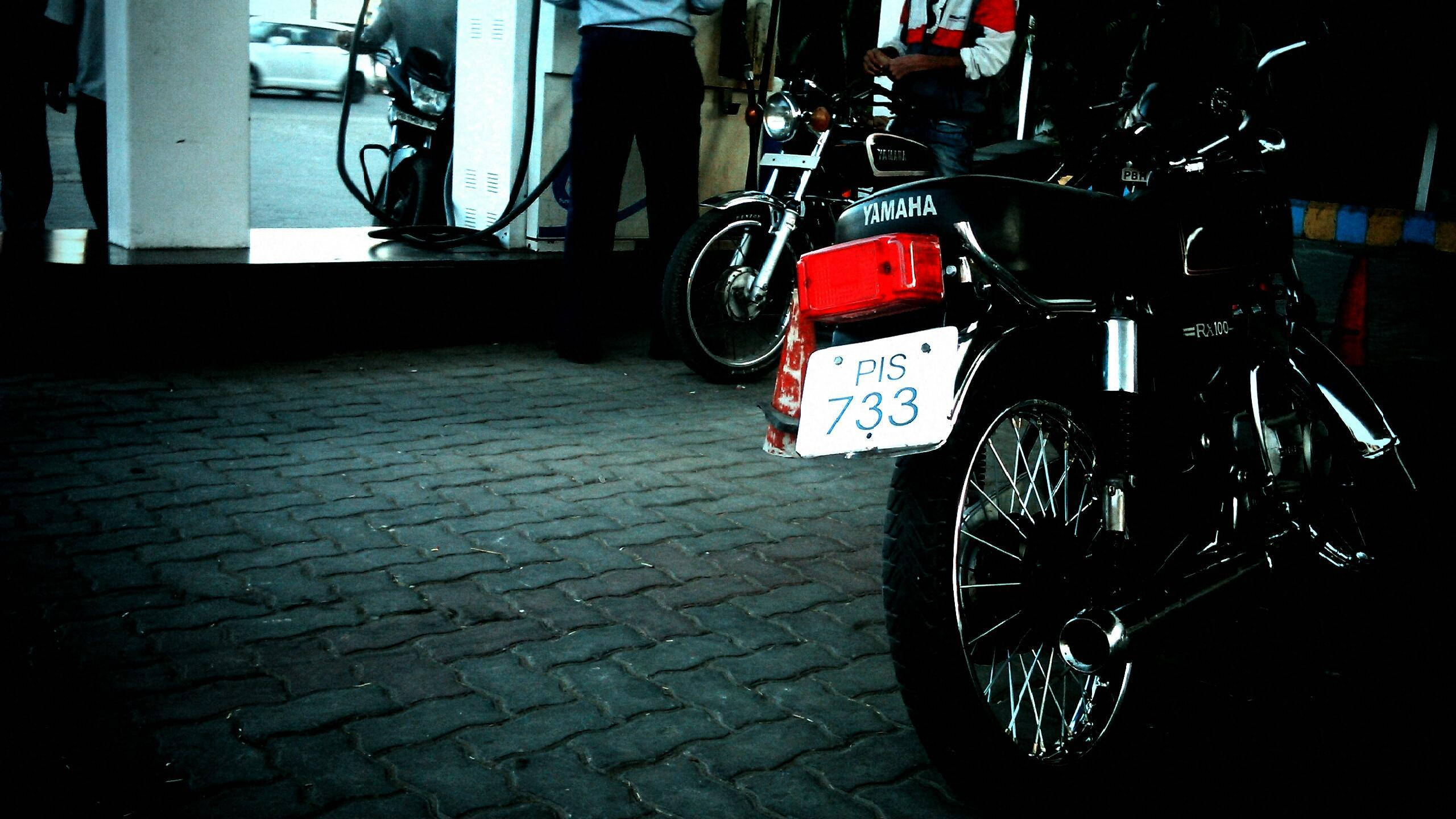 雅馬哈RX100摩托車後部特寫 壁紙 — 一輛黑色Yamaha電單車，車牌PIS 733，夜晚停泊在鵝卵石街道上，燈光昏暗。