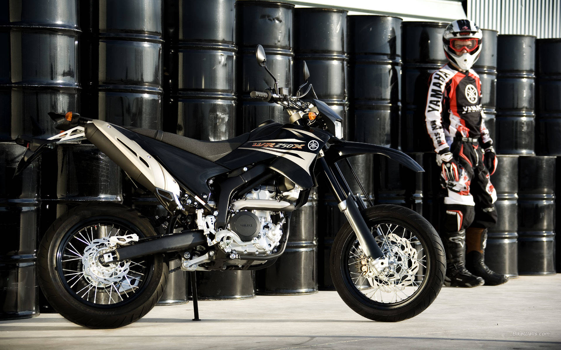 雅馬哈黑色摩托車最佳桌面 壁紙 — 一架黑色和銀色的Yamaha越野電單車用側腳架支撐著，旁邊站著一位全副裝備的騎手，背景是堆疊的黑色油桶。