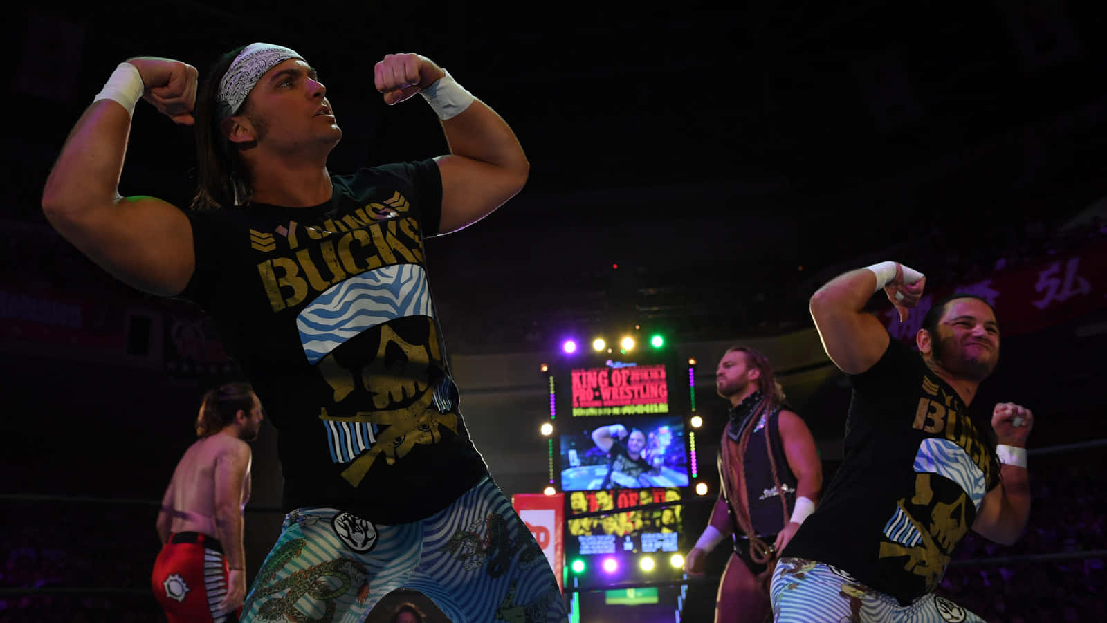 雙人組合Matt與Nick Jackson，即The Young Bucks 壁紙 — 兩名肌肉發達的摔角手在燈光璀璨的擂台上勝利地展示肌肉，背景有色彩繽紛的舞台燈光和人群。