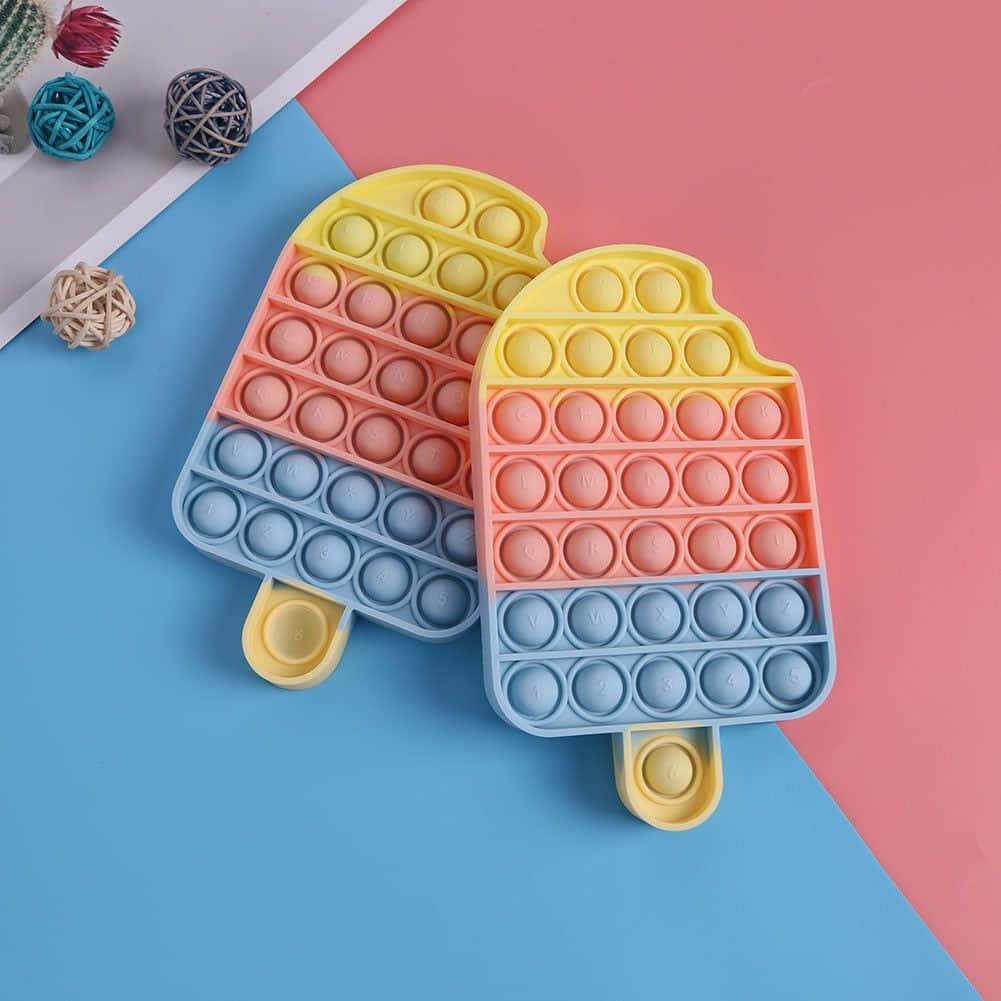 雙冰淇淋Pop It玩具圖 壁紙 — 兩個色彩繽紛的泡泡減壓玩具，形狀像冰棒，放在充滿活力的藍色和粉紅色幾何背景上。