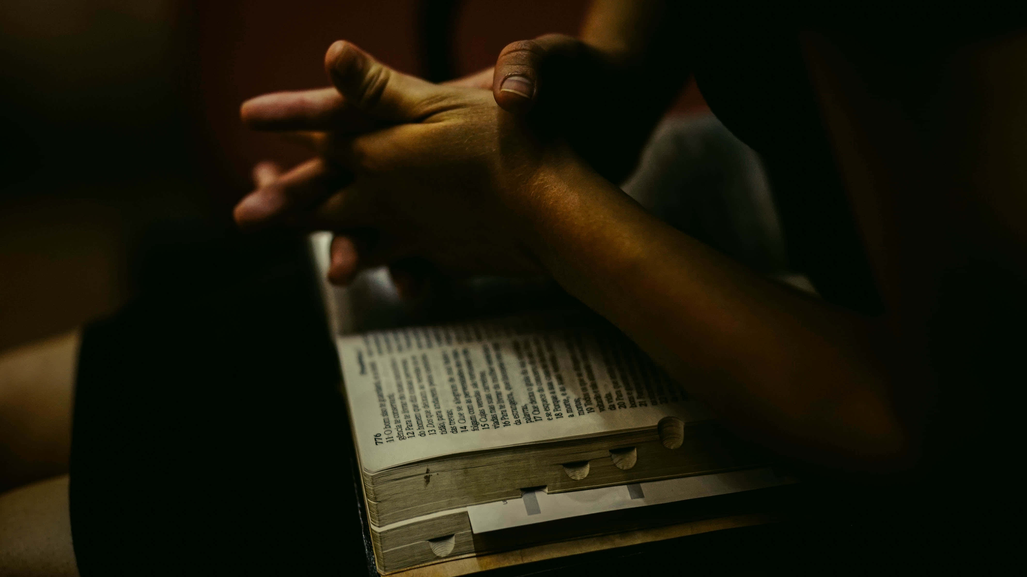 Gospel 桌布 壁紙 — 雙手交疊於一本打開的書上，在昏暗溫暖的光線中，營造出靜謐沉思的時刻。