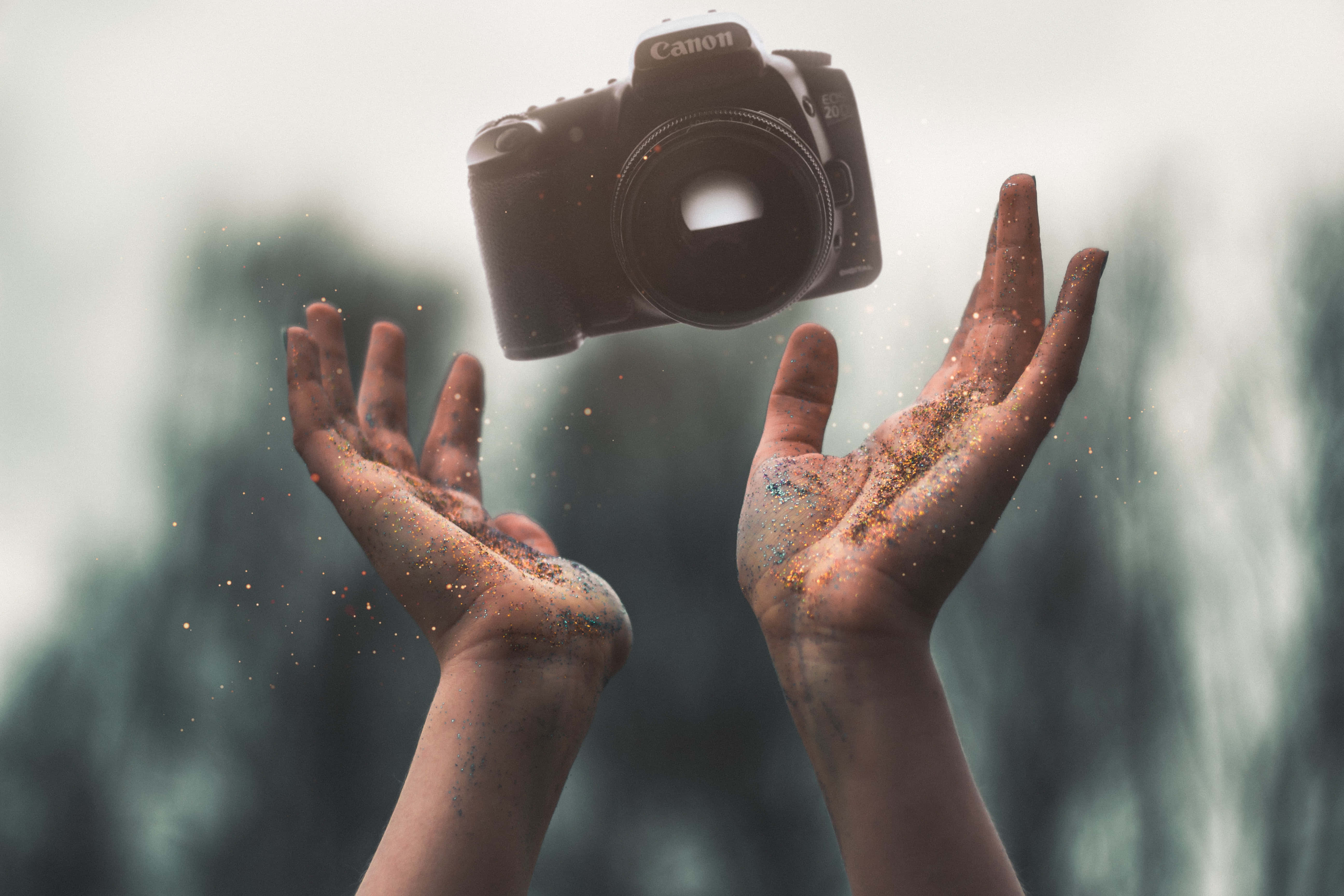 hands 桌布 壁紙 — 雙手向上捧起，閃爍的粒子在周圍漂浮，一部黑色Canon相機懸浮在上方。