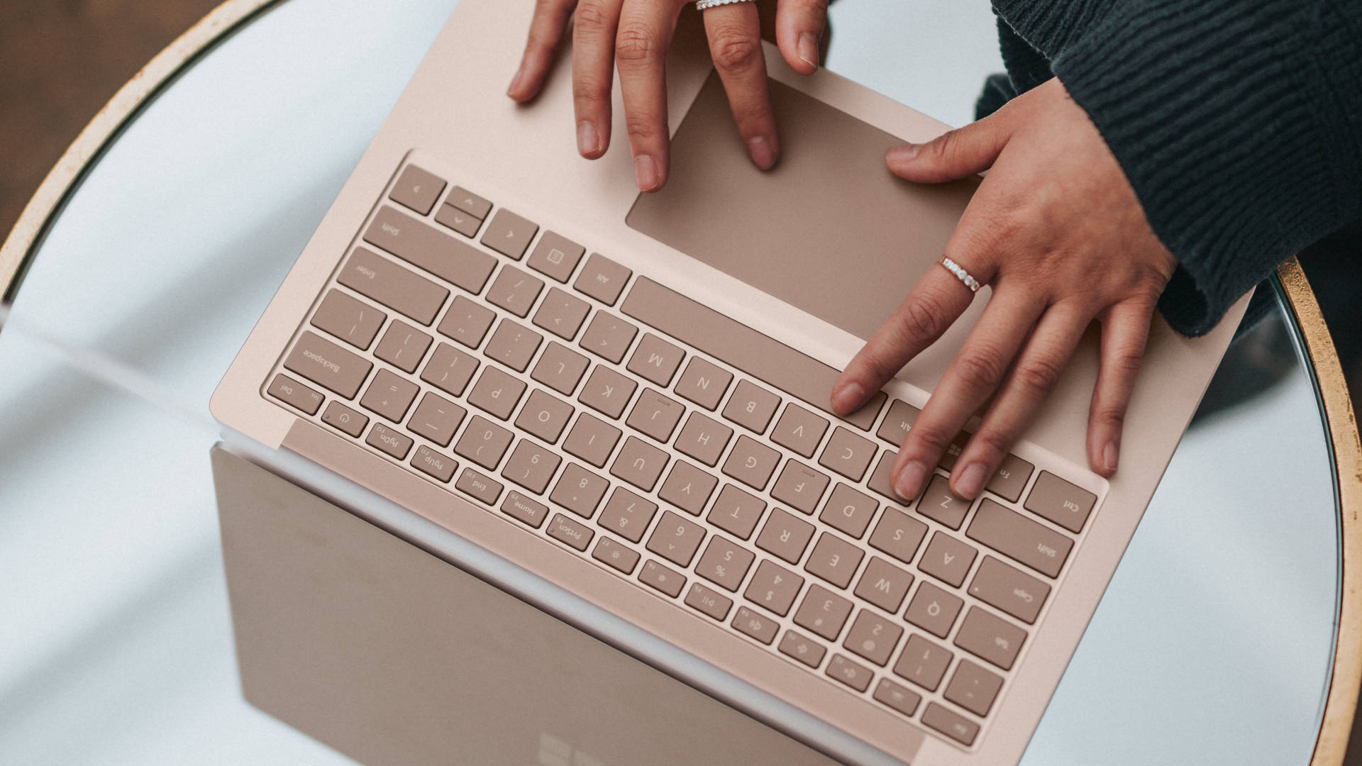 microsoft 桌布 壁紙 — 雙手在玫瑰金色筆記型電腦上打字的特寫，電腦放在帶木邊的玻璃桌上。