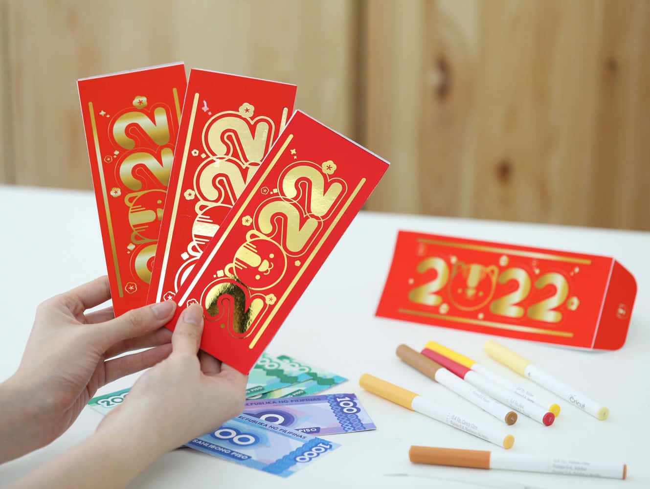 Red Envelope 桌布 壁紙 — 雙手在白色桌子上，拿著帶有金色2022設計的紅色利是封，放在貨幣和記號筆上方。