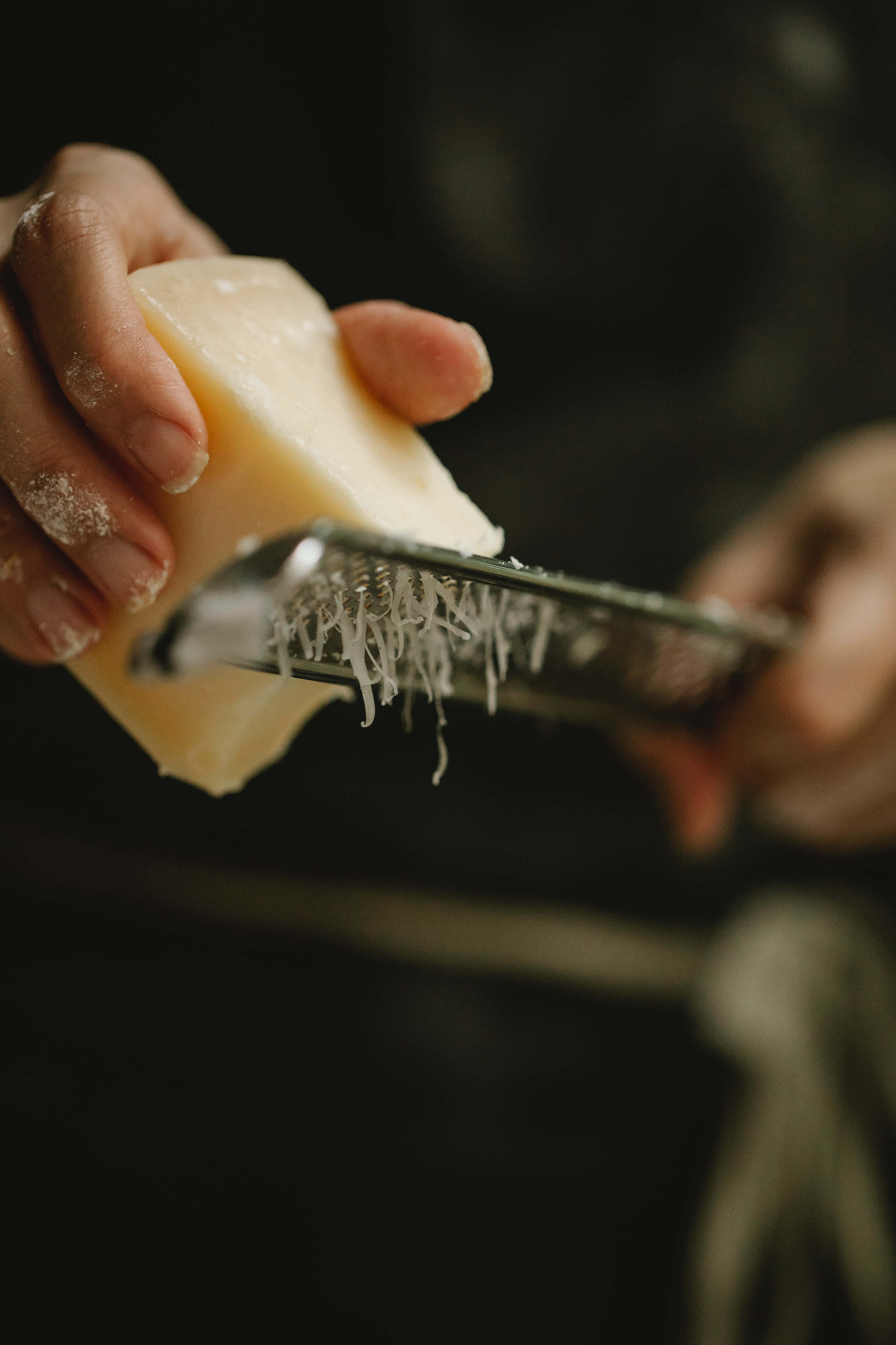Cheese 桌布 壁紙 — 雙手將淡色芝士塊在金屬刨絲器上刨絲的特寫，細碎的芝士絲落下。
