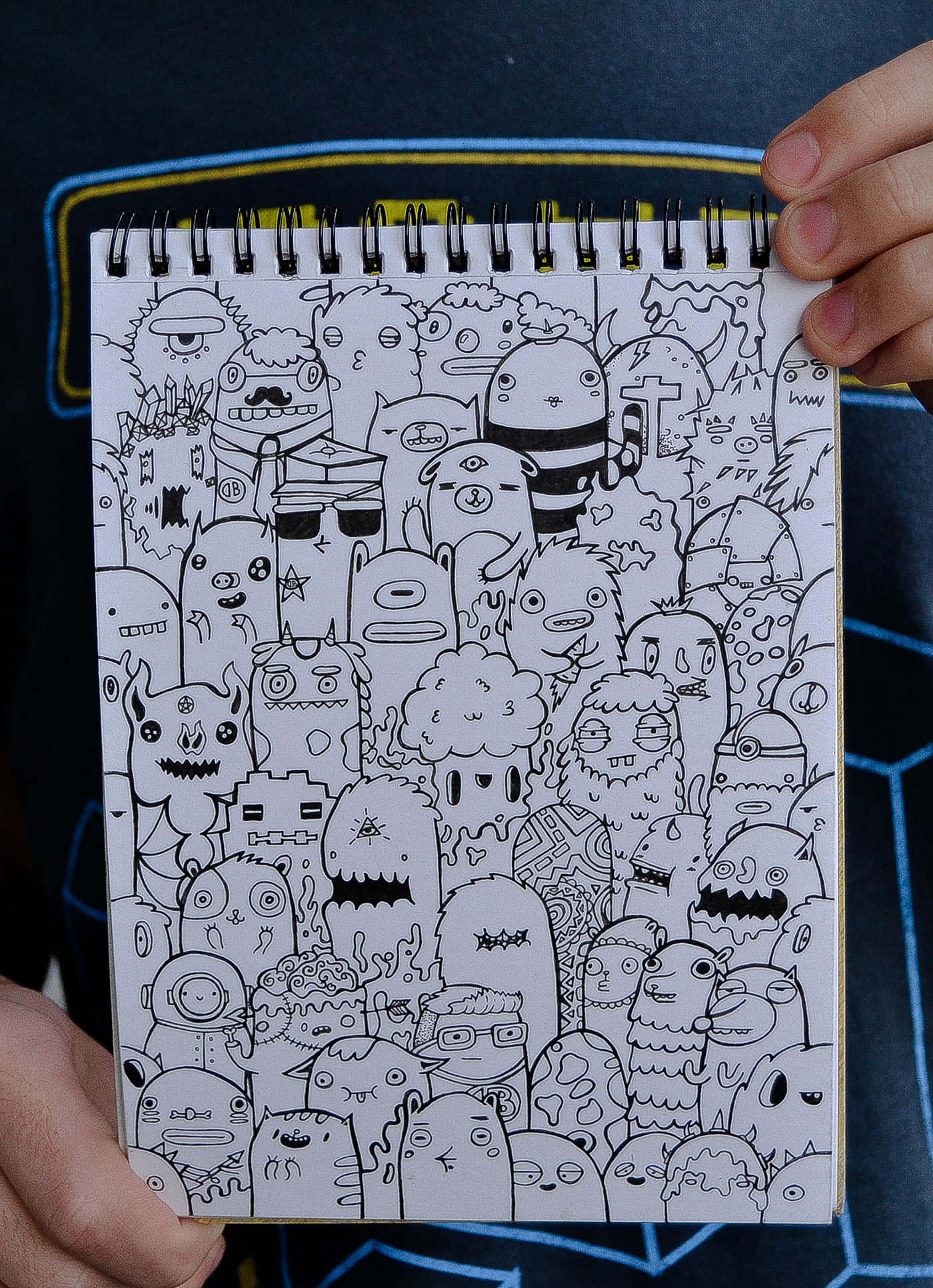 塗鴉 桌布 壁紙 — 雙手拿著一本素描簿，裡面密密麻麻畫滿了用黑色墨水畫在白紙上的古怪卡通怪物。