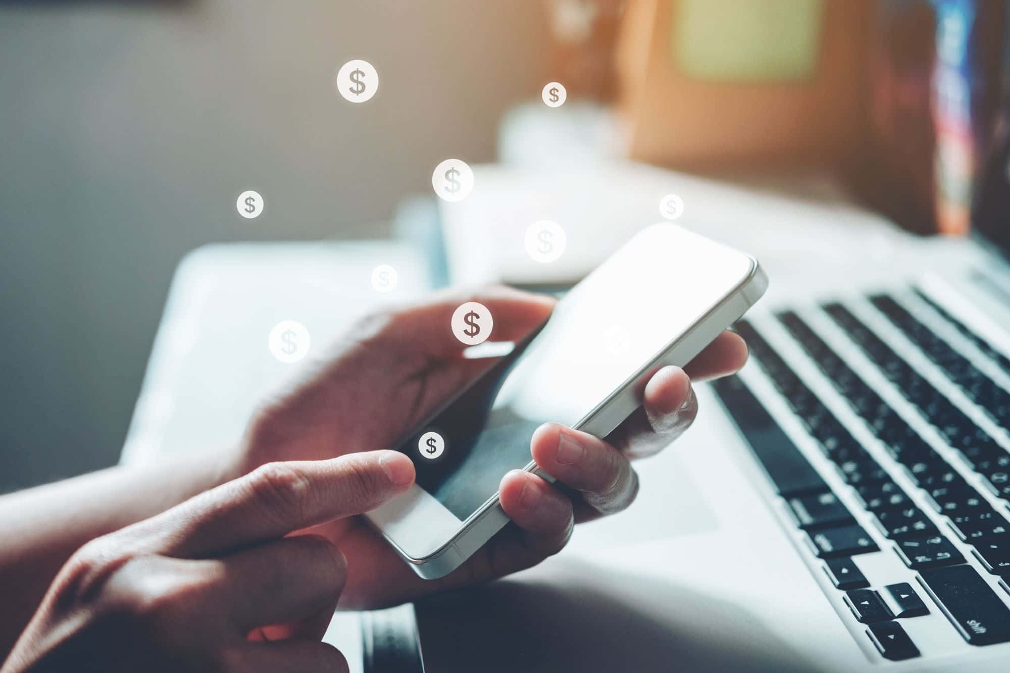 Mobile Banking 桌布 壁紙 — 雙手握住一部智能手機，上面漂浮著美元符號，旁邊有一部手提電腦，在柔和光線的工作空間中。