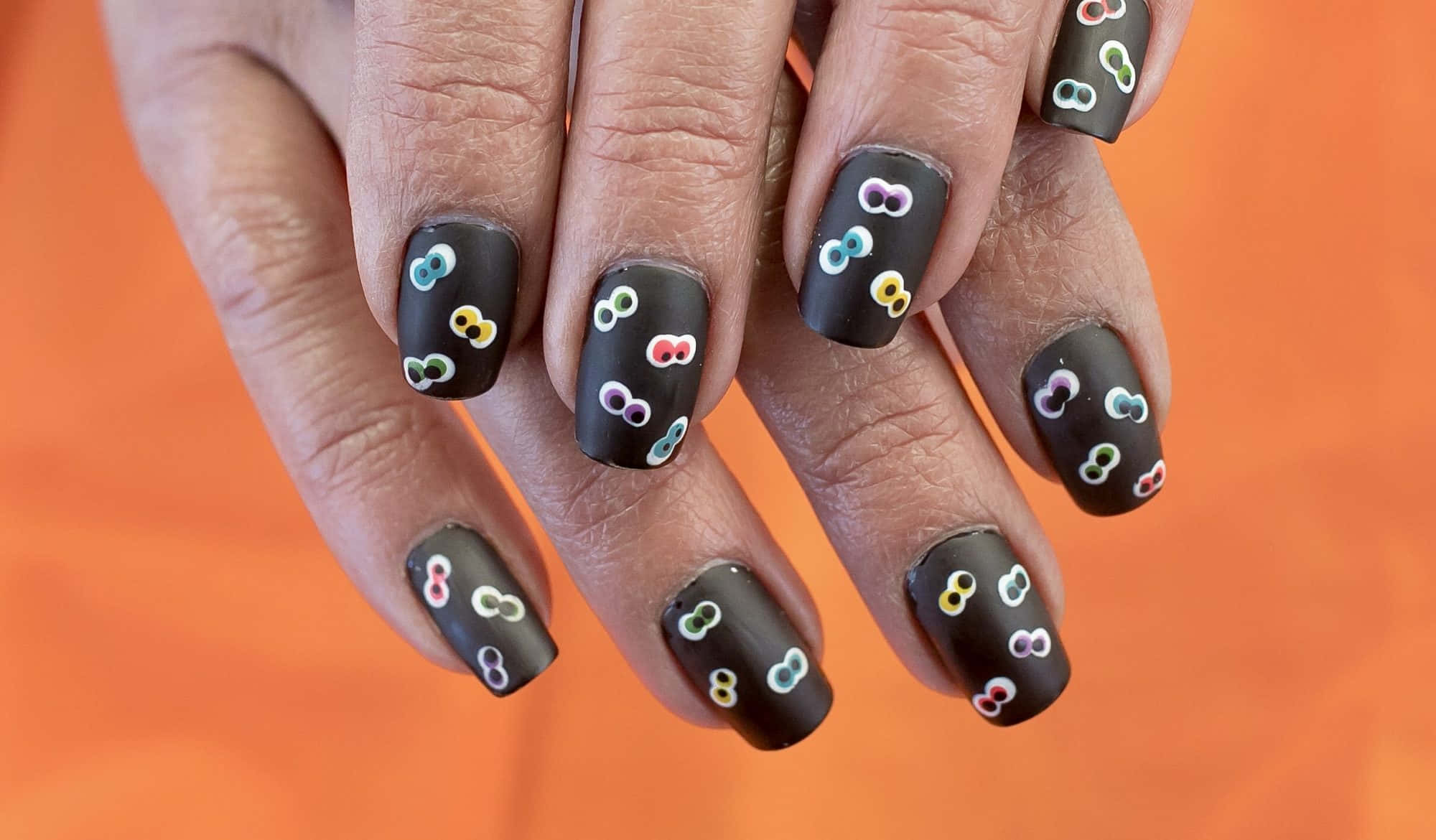 Halloween Nail Art 桌布 壁紙 — 雙手特寫，黑色啞光指甲上飾有色彩繽紛的小無限符號，背景為鮮豔的橙色。