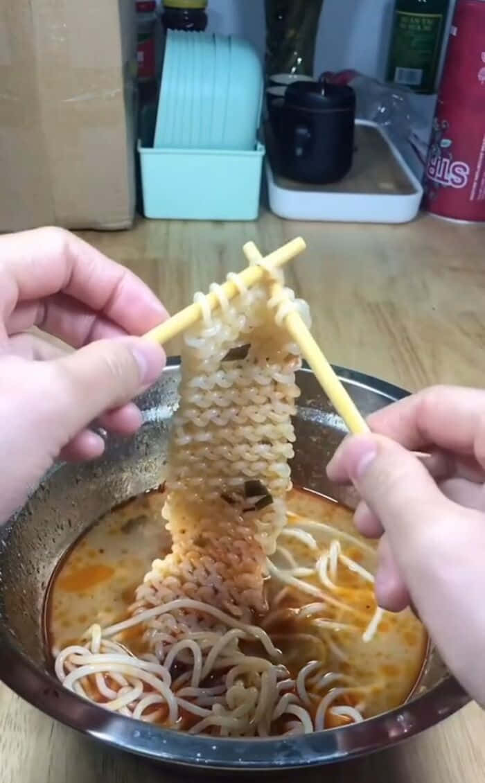 Food Memes 桌布 壁紙 — 雙手用筷子從金屬碗中夾起冒煙辣味拉麵的特寫。