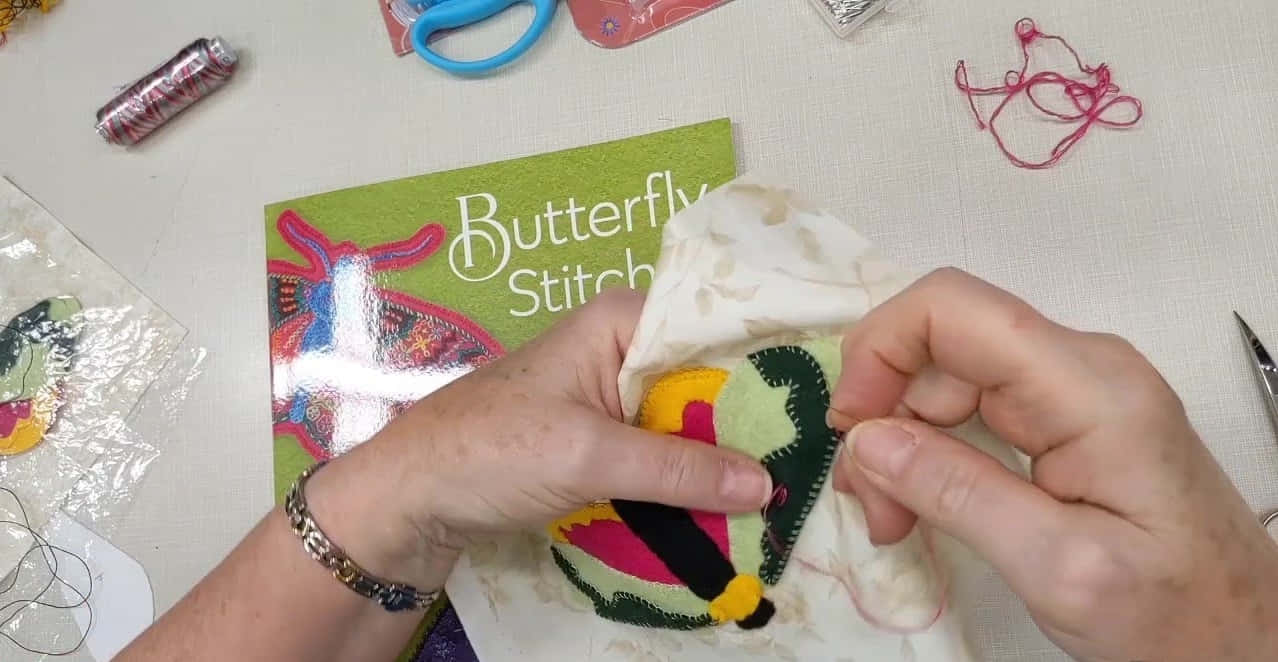 Butterfly Stitch 桌布 壁紙 — 雙手用紅線小心地將彩色蝴蝶貼花縫在布料上，周圍是手工藝用品。