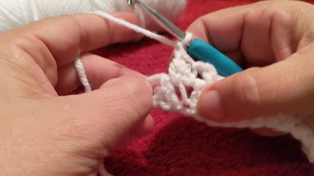 Butterfly Stitch 桌布 壁紙 — 雙手用青綠色鉤針編織白色紗線的特寫，背景為紅色紋理背景。