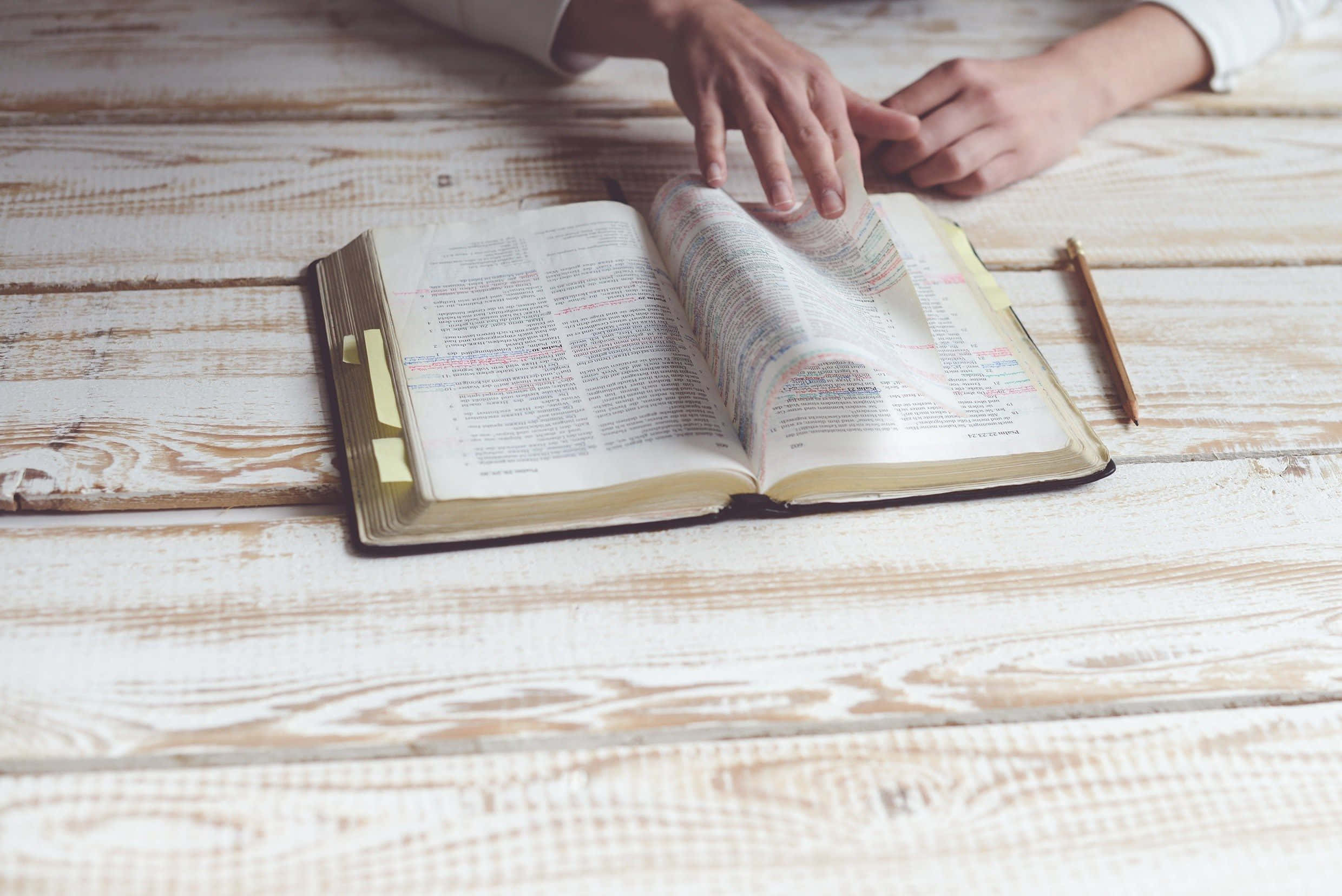 Bible Study 桌布 壁紙 — 雙手翻開一本破舊聖經的頁面，放在白色粉刷的木桌上，旁邊有一支鉛筆。