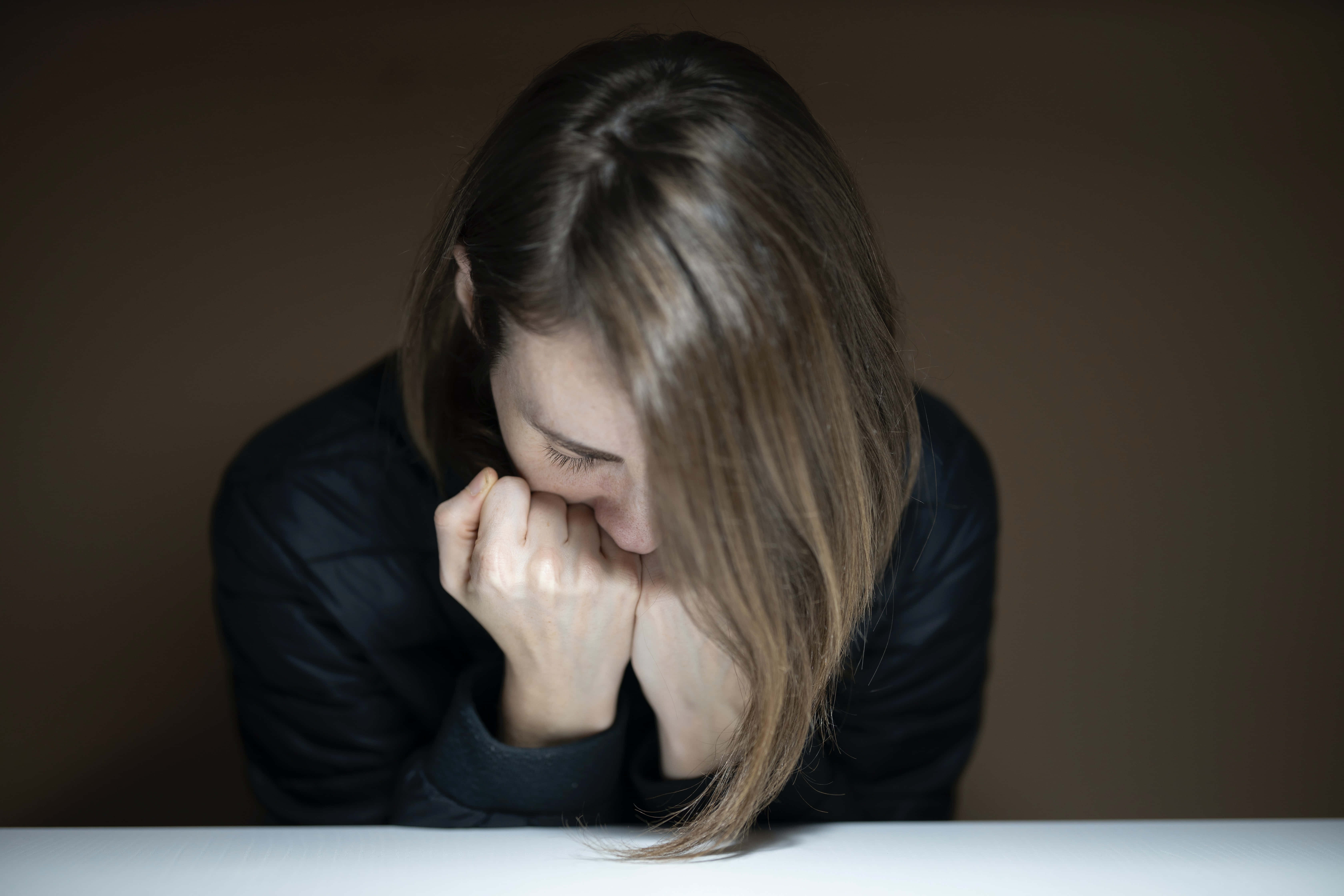 雙拳抵臉的憂鬱女子圖 壁紙 — 一位長長啡色頭髮的女性在燈光昏暗的房間裡，將頭靠在白色桌子上緊握的雙手上。