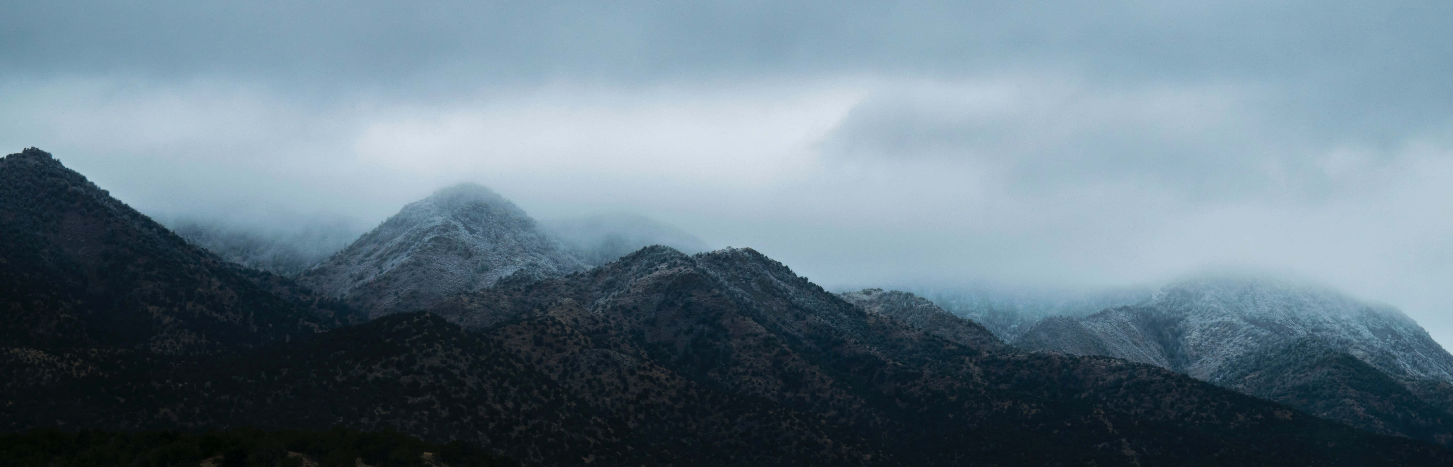 雙螢幕自然風景 壁紙 — 灰色陰天下部分被雪覆蓋的霧山，帶有陰暗的森林斜坡。