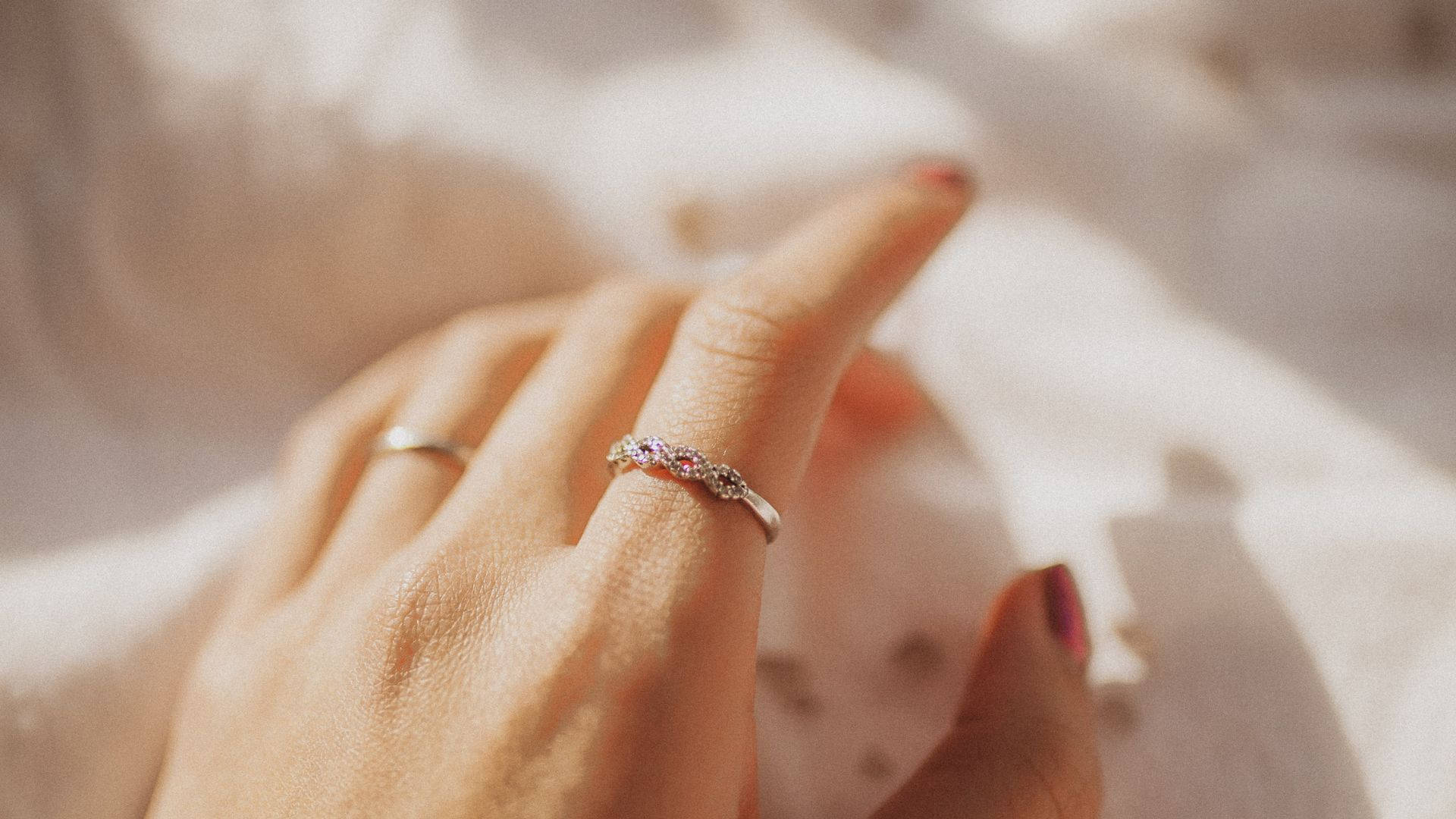 雙鑽扭結婚戒 壁紙 — 一隻手戴著兩枚戒指的特寫，其中一枚飾有粉紅色和透明寶石，放在柔軟的白色布料上。
