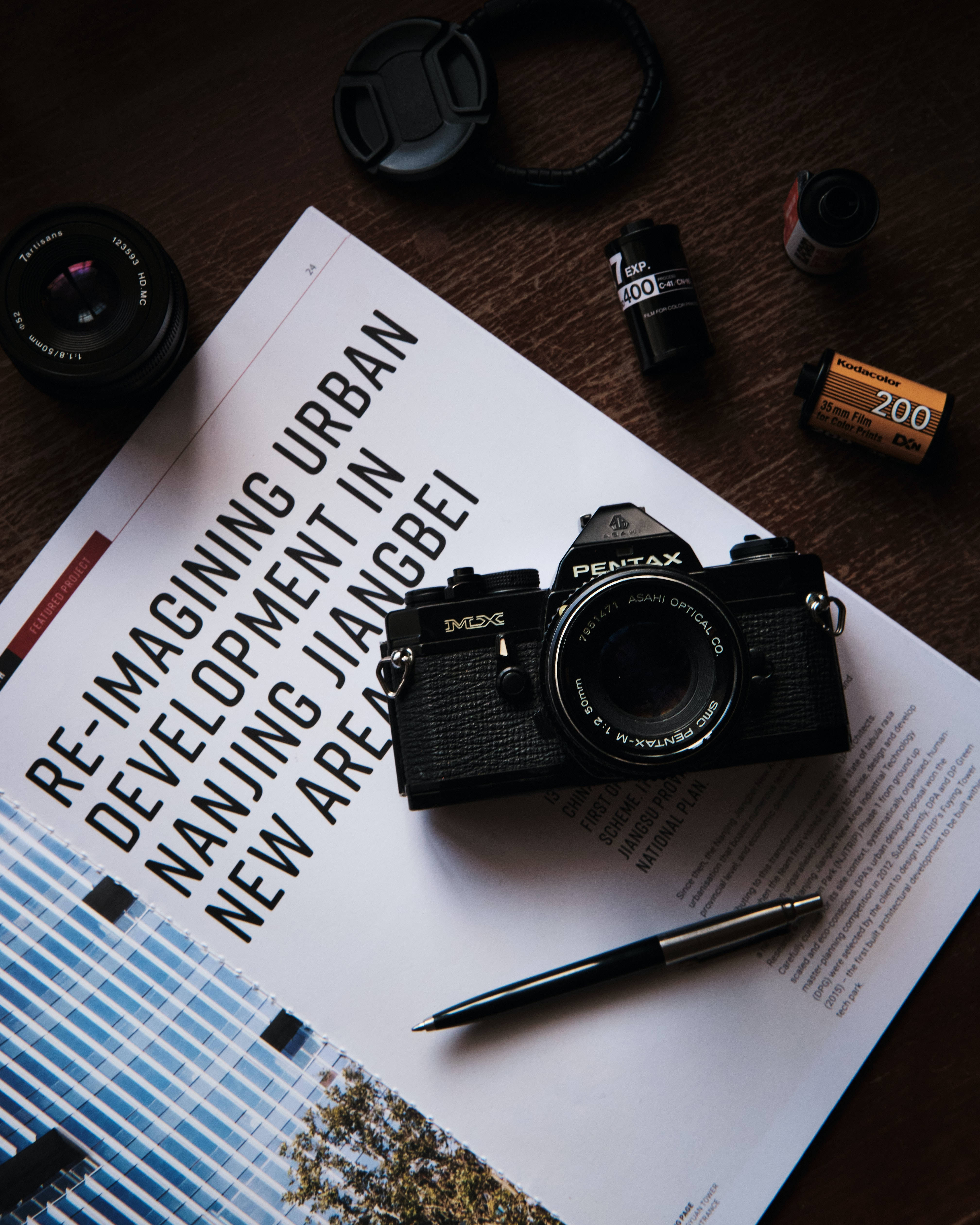 雜誌與老式相機 壁紙 — 一部復古黑色Pentax菲林相機放在一本關於城市發展的雜誌上，周圍有鏡頭和菲林卷在木質表面上。