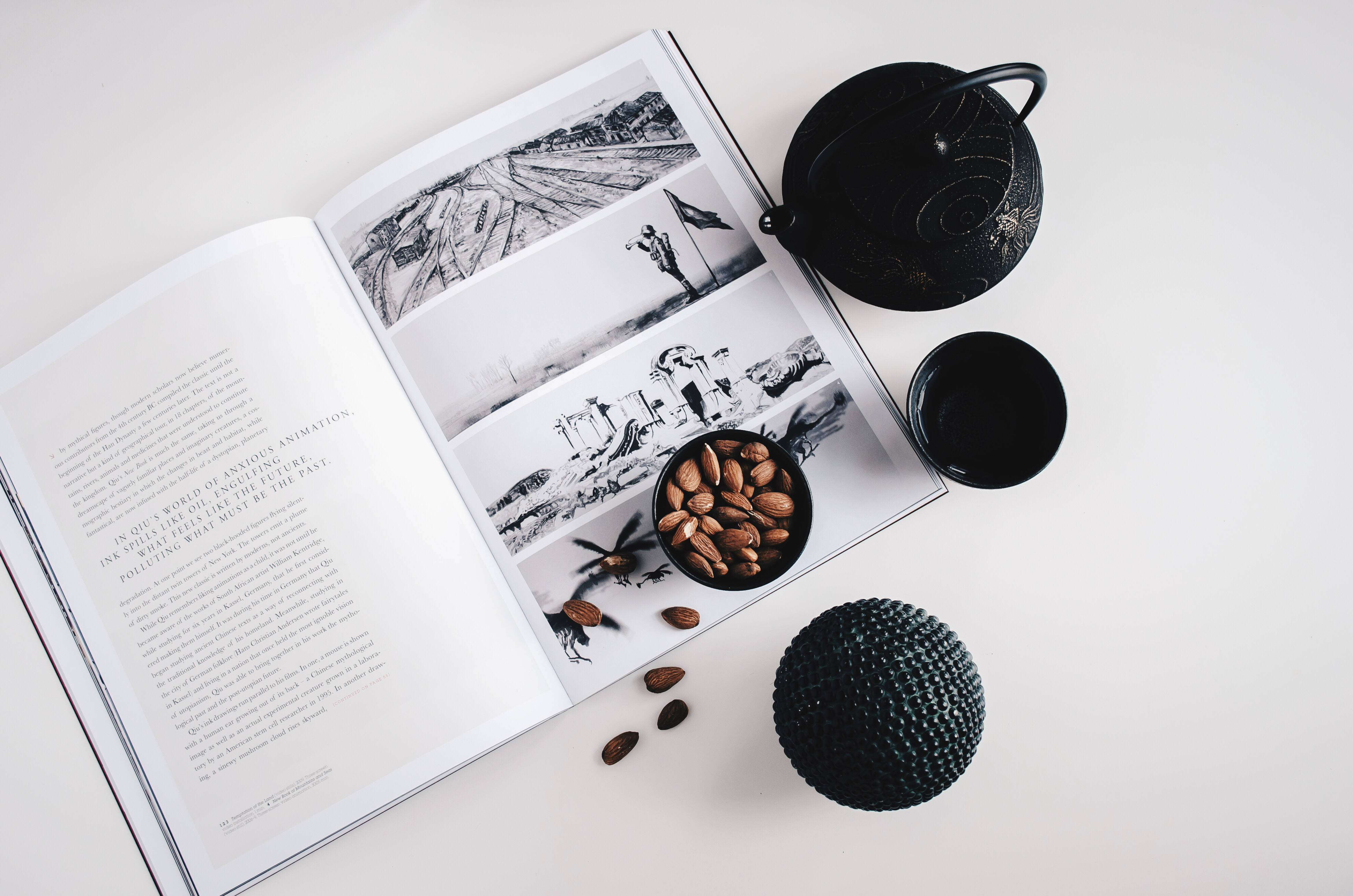 雜誌與黑色陶瓷器 壁紙 — 一個極簡風格的平鋪構圖，包含一本打開的書，書中有黑白插圖，一個黑色茶壺、杯子以及杏仁，排列在白色表面上。
