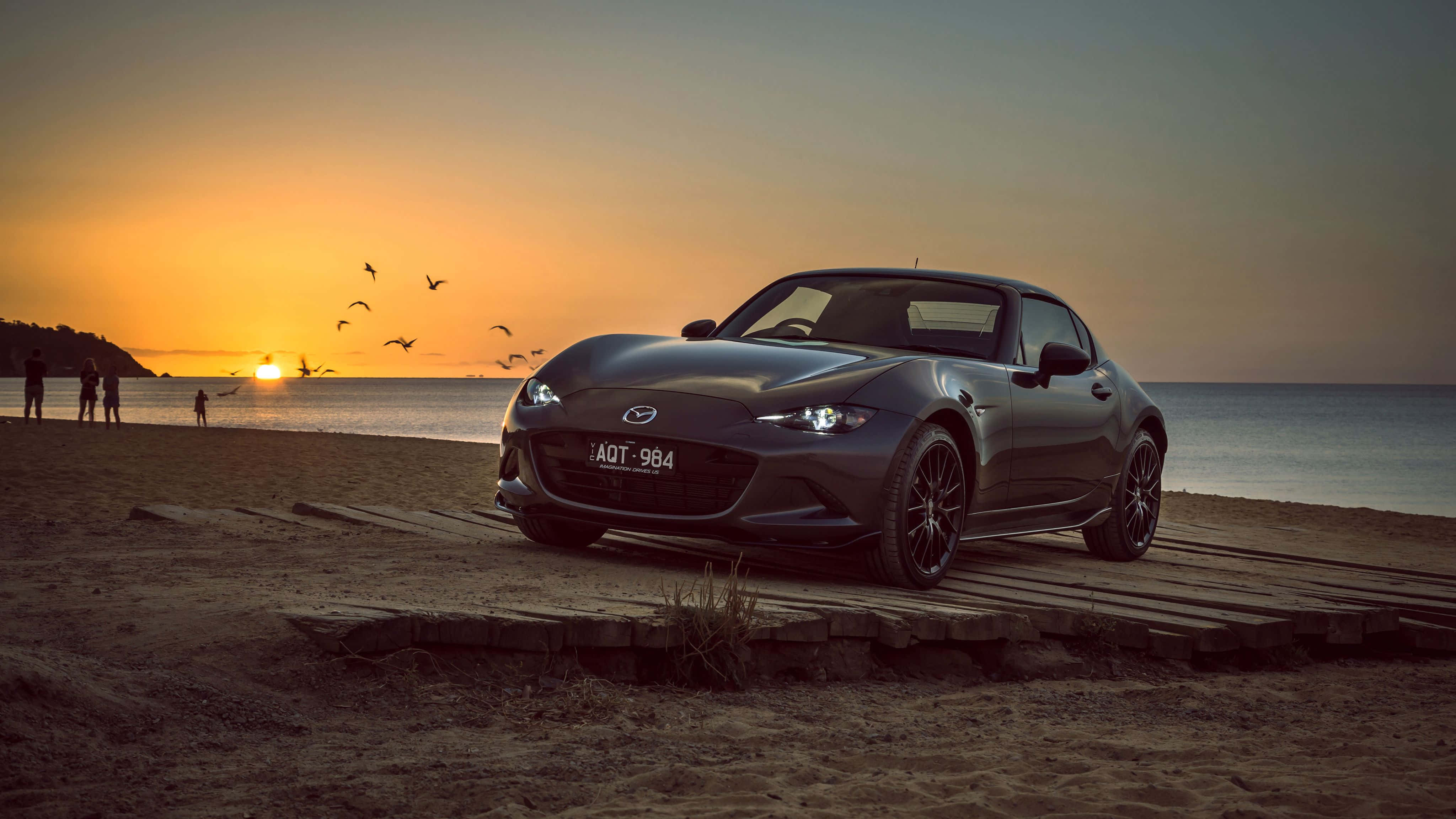 難忘的時尚：經典馬自達MX-5 Miata 壁紙 — 一架時尚深灰色萬事得MX-5 Miata停泊在日落時分的木製木板路上，背景有剪影的海灘遊客和飛鳥。