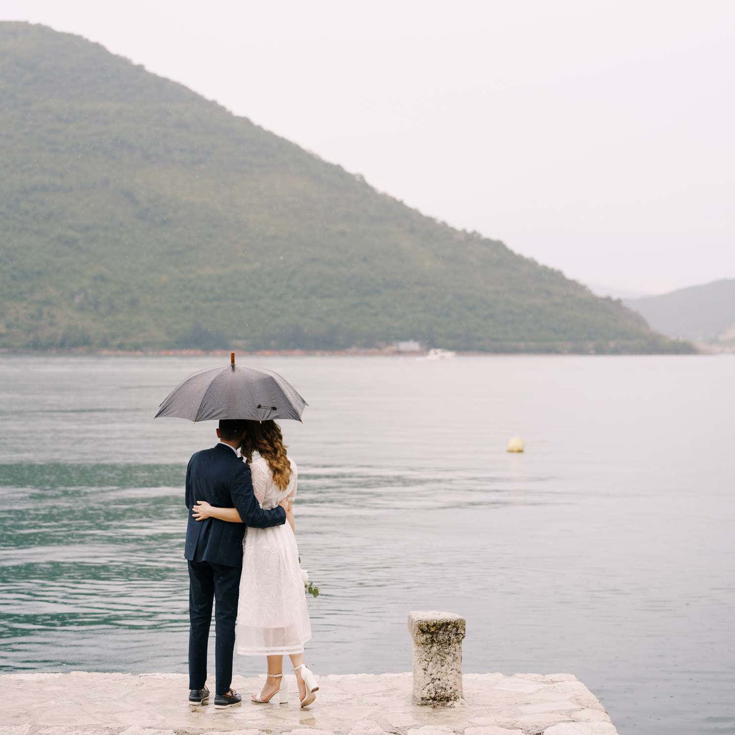 雨中兩位戀人共享美好時光 壁紙 — 一對情侶在石碼頭上撐著灰色雨傘擁抱，俯瞰平靜的大海，背景是霧氣瀰漫的綠色山丘。
