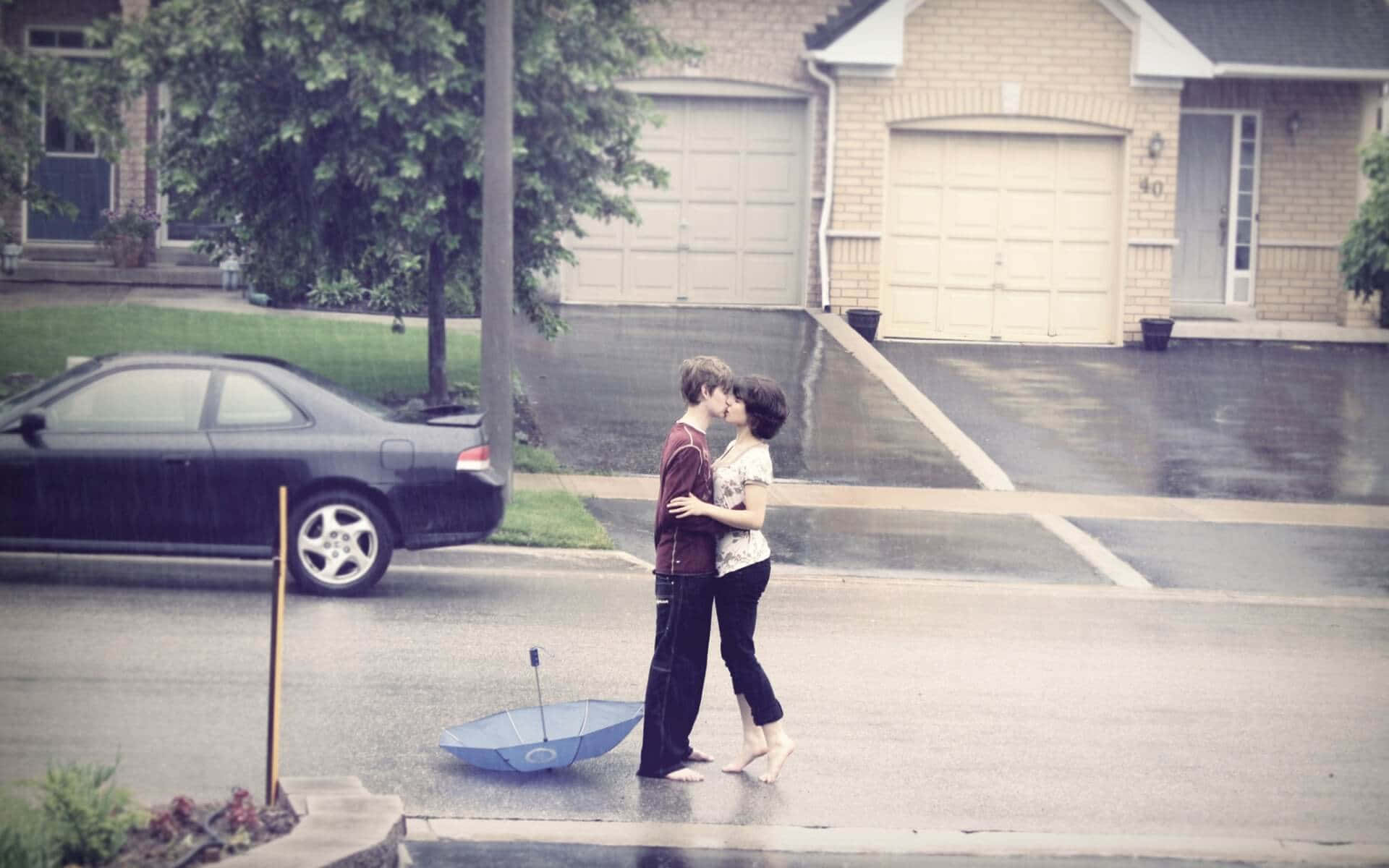 雨中年輕情侶親吻圖 壁紙 — 一對情侶在雨中激情接吻，濕漉漉的車道上有一把藍色雨傘被遺棄在附近。