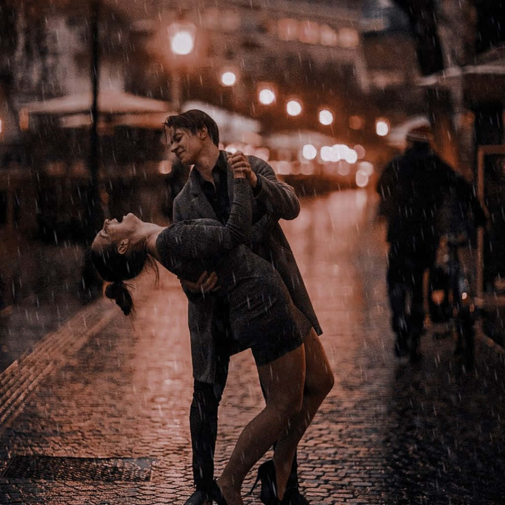 雨中情侶的浪漫時刻 壁紙 — 一對情侶在夜晚溫暖燈光照亮的鵝卵石街道上，在雨中熱情擁抱和跳舞。