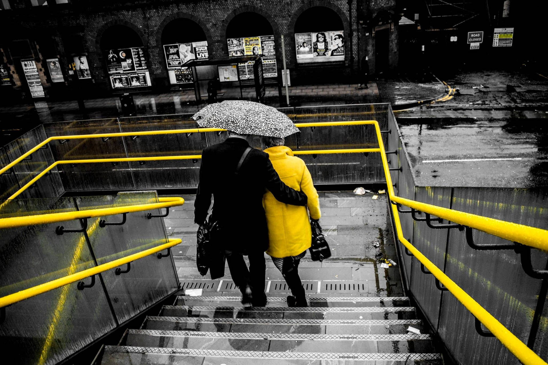 兩人在下雨的城市中撐著雨傘走下濕漉漉的樓梯，樓梯有黃色欄杆。