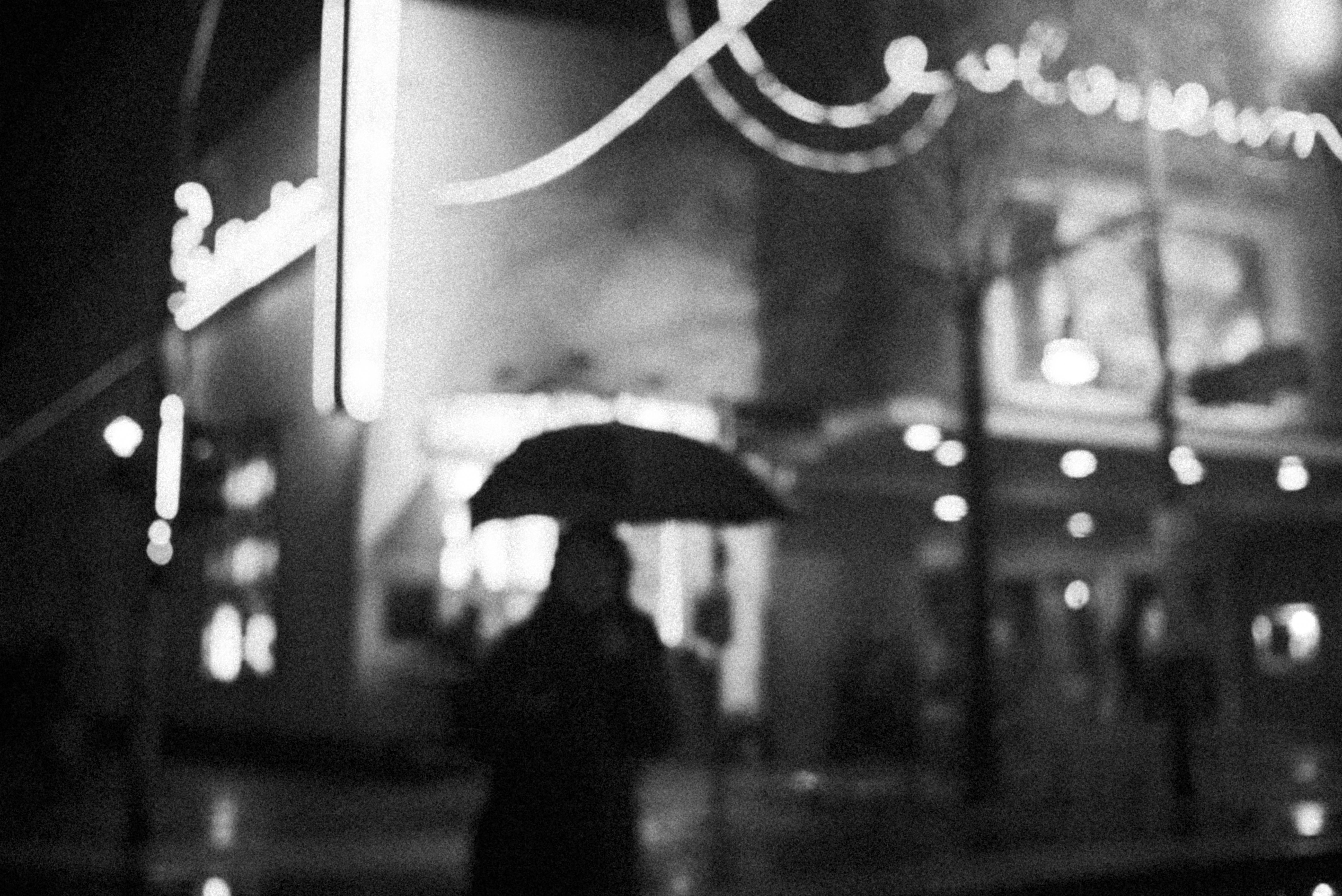 雨中持傘的暗黑憂鬱少女 壁紙 — 一個模糊的人影撐著雨傘，在夜晚閃爍的霓虹燈下走過雨中的城市街道。