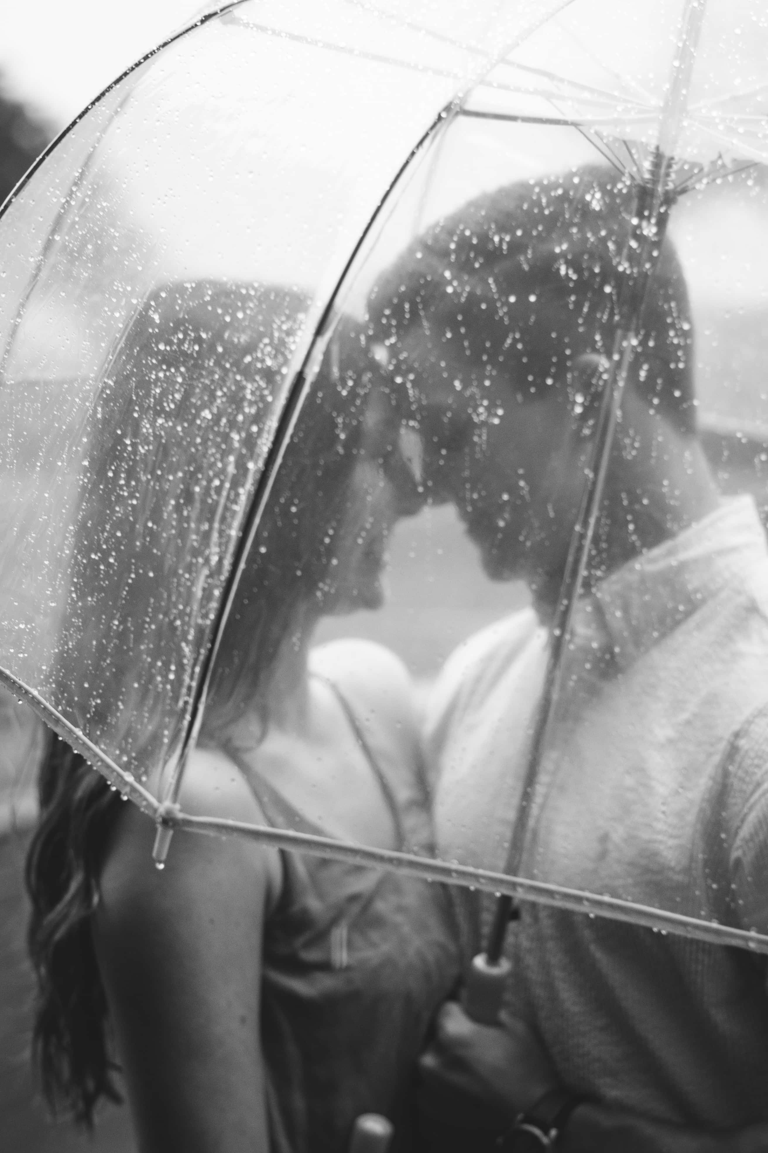 雨中擁抱的兩位戀人 壁紙 — 一張黑白照片，一對情侶在佈滿雨滴的透明雨傘下接吻。