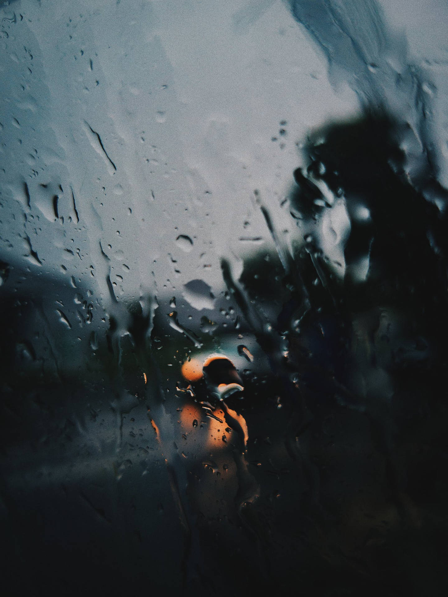 透過濺滿雨水的窗戶看到一個人的剪影，背後有橙色的光。