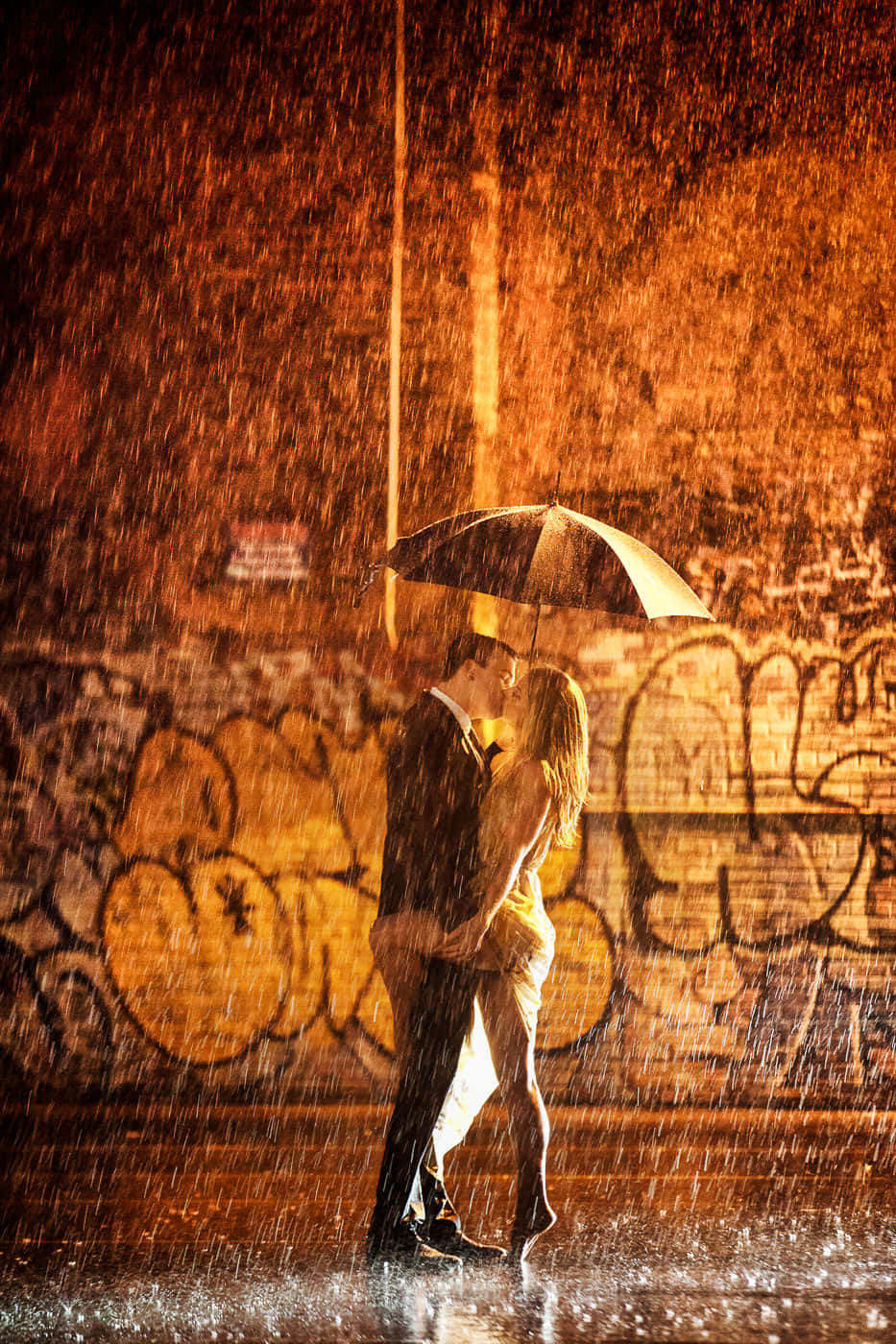 雨中牽手漫步，情侶沉浸於浪漫氛圍 壁紙 — 一對情侶在傾盆大雨中於傘下接吻，背景是塗鴉牆，沐浴在溫暖的橙色燈光中。