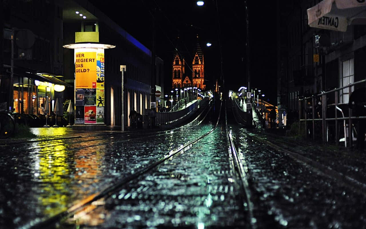 夜間城市街道上的雨，有電車軌道和遠處燈火通明的教堂。
