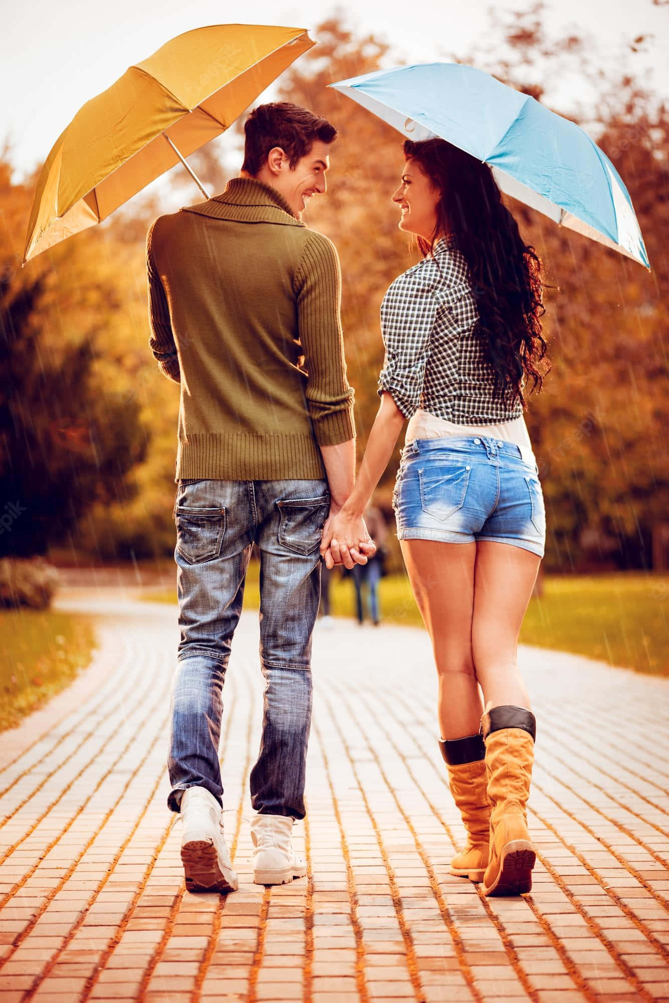 雨中雙傘情侶圖 壁紙 — 一對微笑的情侶手牽手走在色彩繽紛的雨傘下，在秋樹環繞的雨中磚路上漫步。