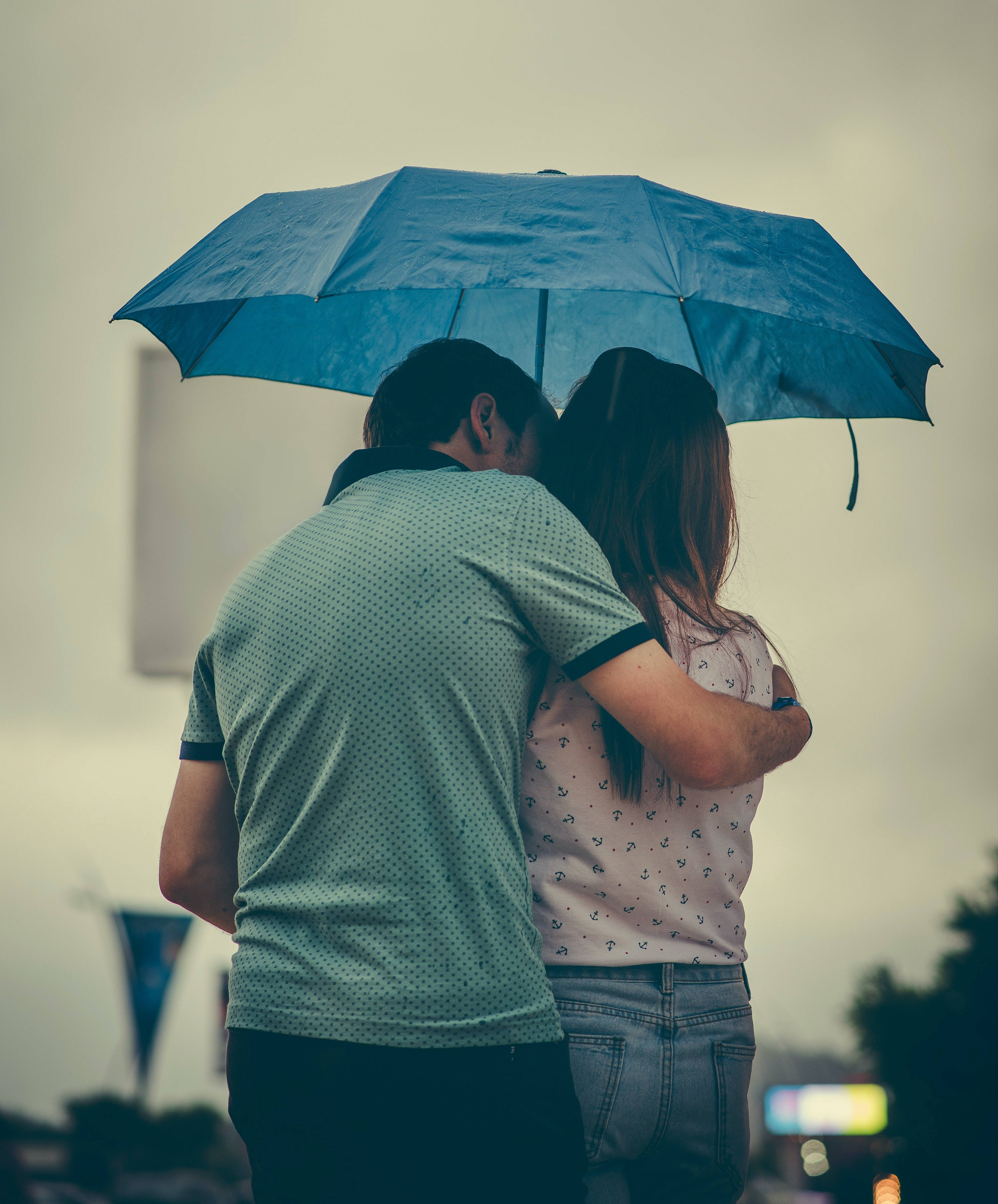 雨傘下擁抱的愛 壁紙 — 一對情侶在雨中藍傘下擁抱，從背後觀看，背景是柔和的城市景觀。