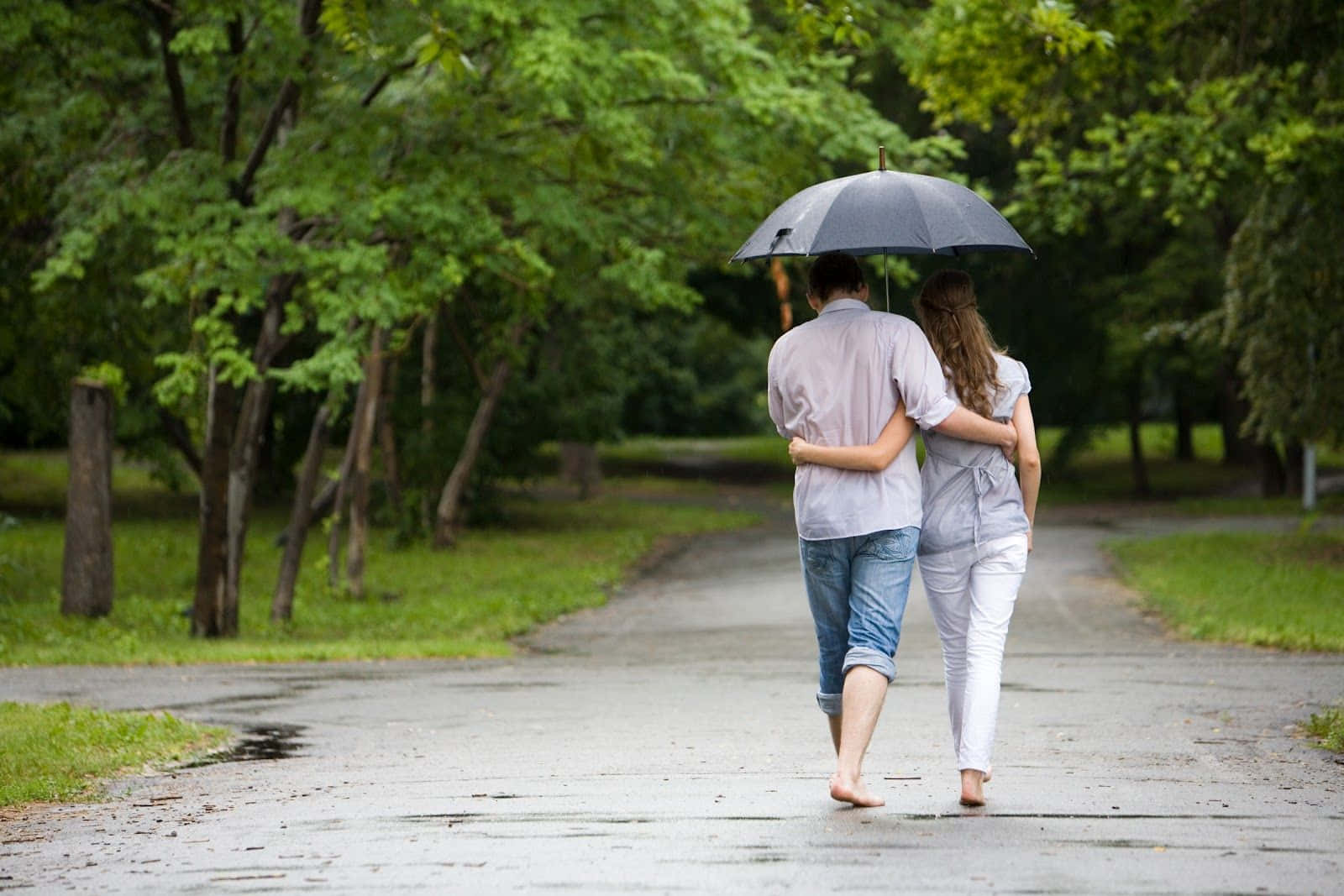 雨傘下的夫妻情侶圖 壁紙 — 一對情侶赤腳撐傘走在潮濕的公園小徑上，四周綠樹成蔭。