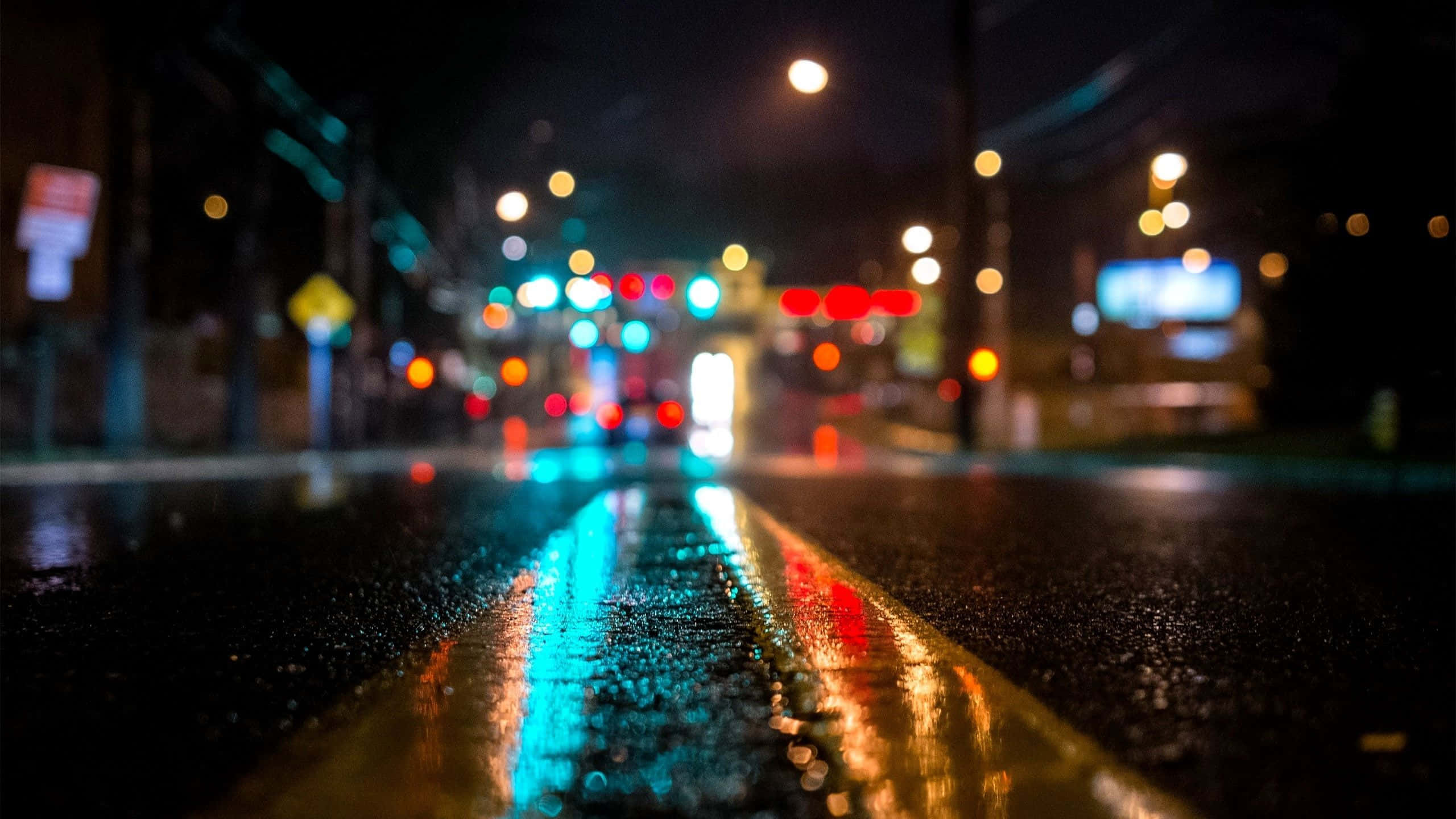 暗夜城市 桌布 壁紙 — 雨夜中的城市街道，濕漉漉的瀝青路面和黃色車道標線反射著閃爍的散景燈光。