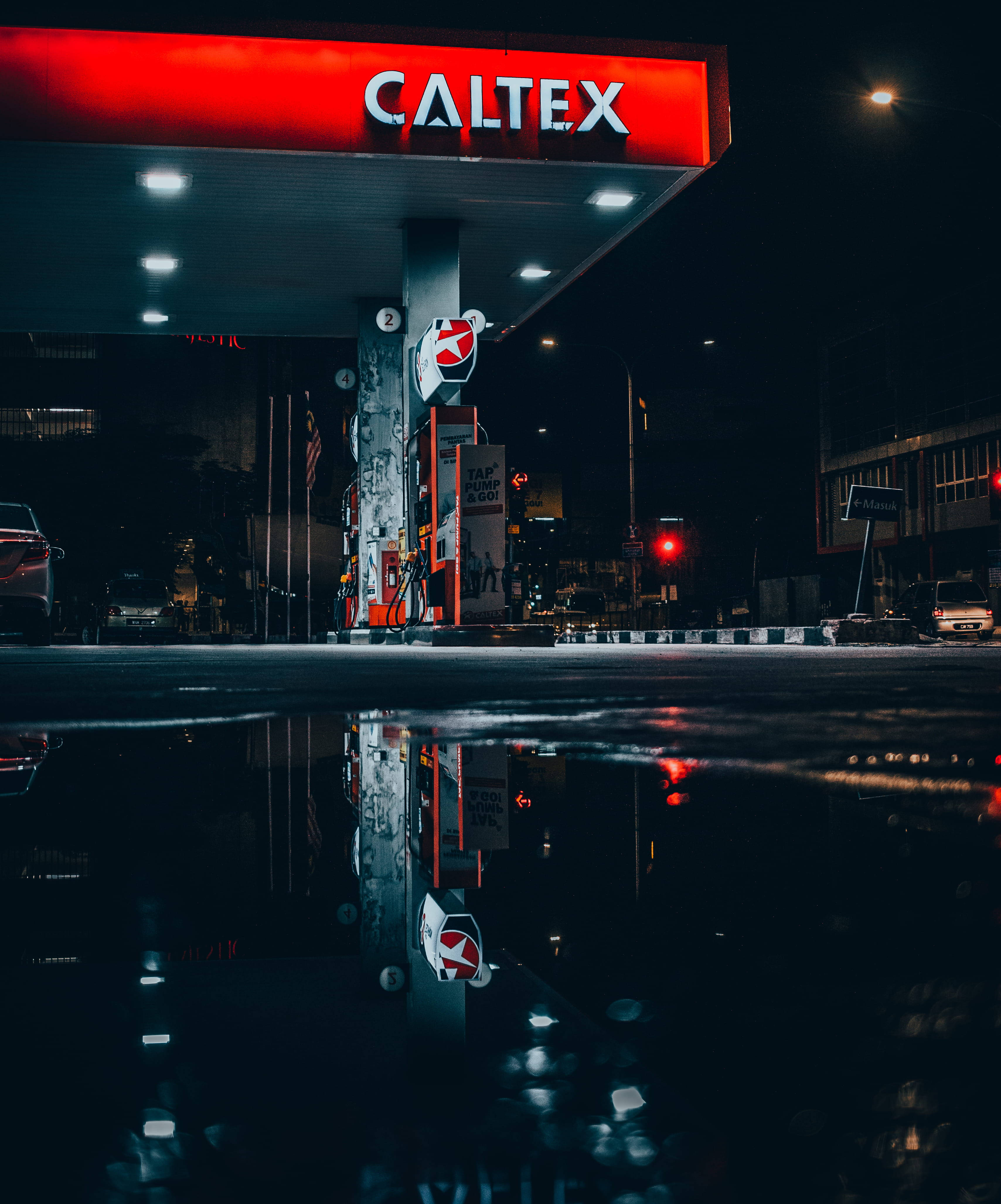 雨夜加德士加油站 壁紙 — 夜晚的Caltex加油站，紅色標誌和油泵倒映在濕漉漉路面上的雨水坑中。