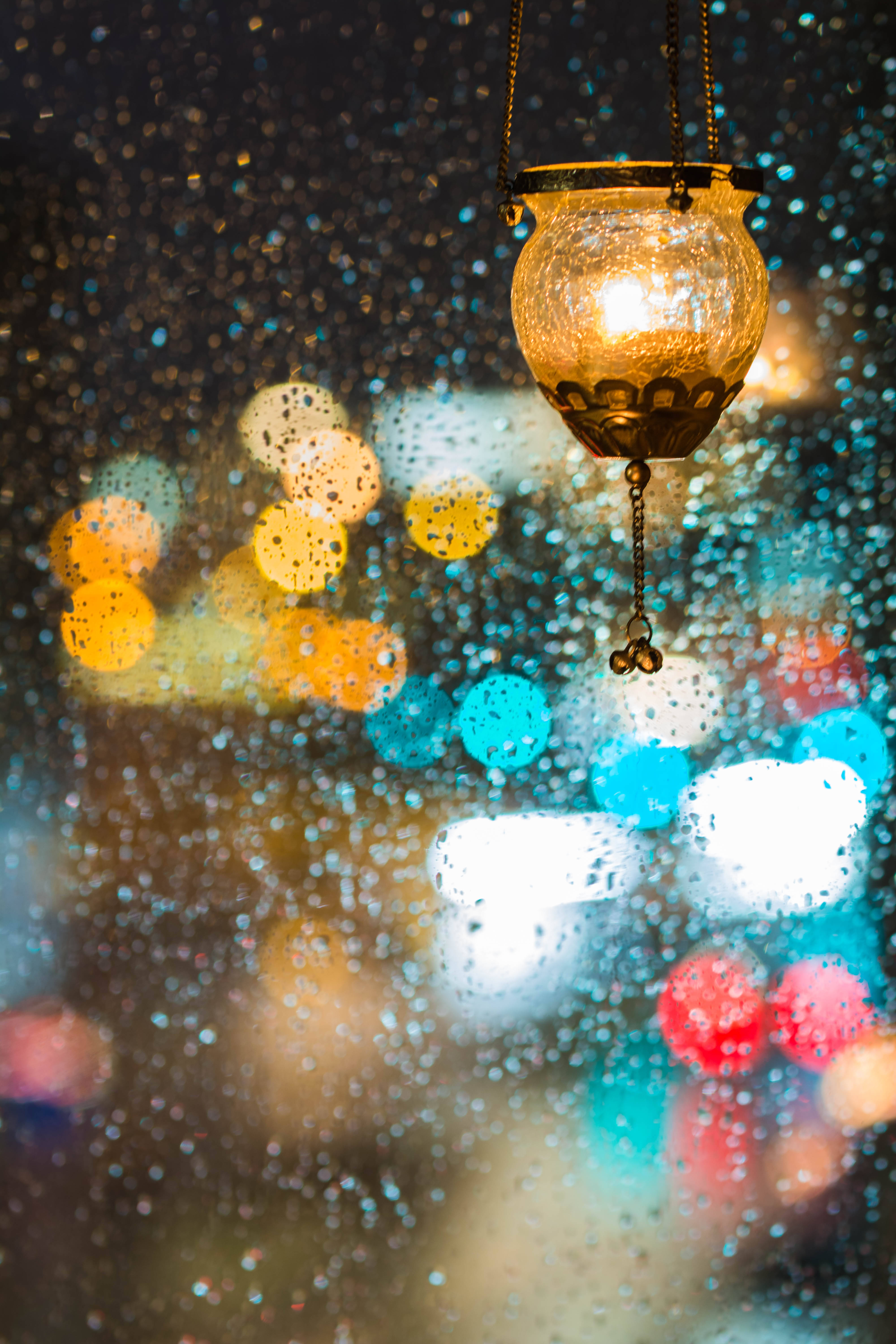 雨夜窗前散景背景 壁紙 — 一盞溫暖發光的燈籠懸掛在佈滿雨點的玻璃後，背景是夜晚充滿活力的散景城市景觀。