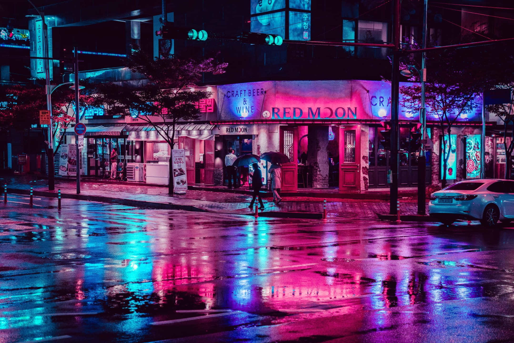 雨夜霓虹城市風景 壁紙 — 雨濕的夜晚城市街道反射商店櫥窗的粉紅和藍色霓虹燈光，行人撐傘走過。