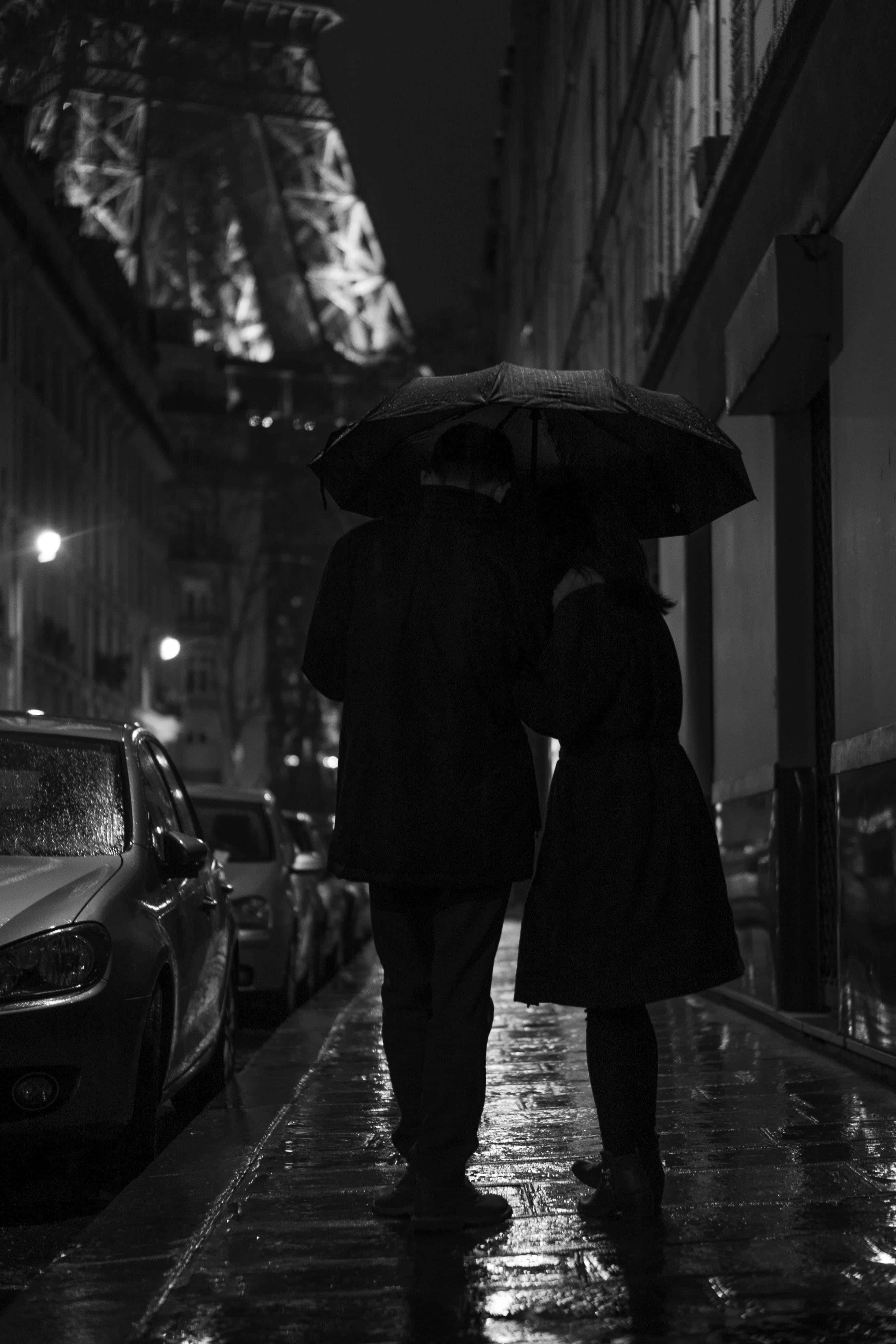 雨天最美的事，是與所愛之人共度 壁紙 — 一對情侶在雨濕的夜晚街道上共用一把傘，遠處的艾菲爾鐵塔閃爍著光芒。