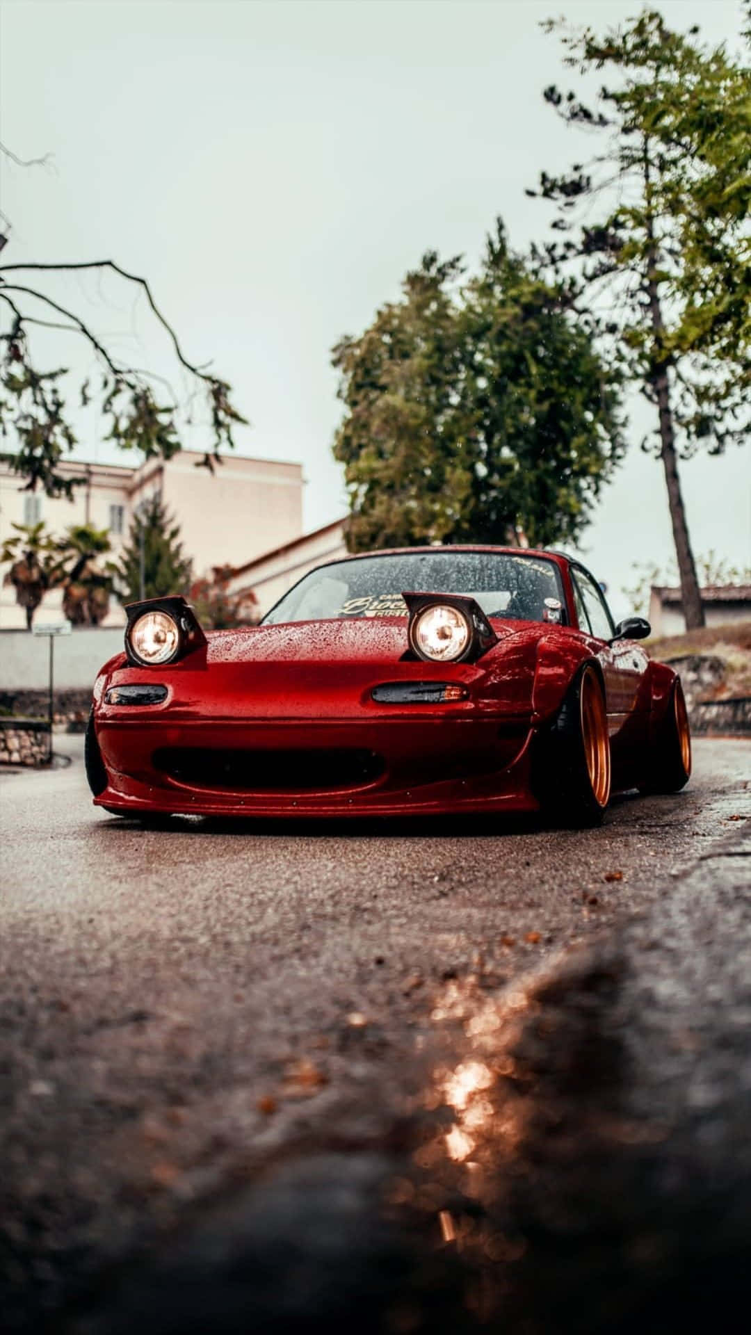 雨後濕潤的1995年馬自達MX-5 Miata紅色跑車 壁紙 — 一輛光澤紅色改裝跑車配金色輪圈，低趴於潮濕路面，背景為陰天、樹木和建築物。