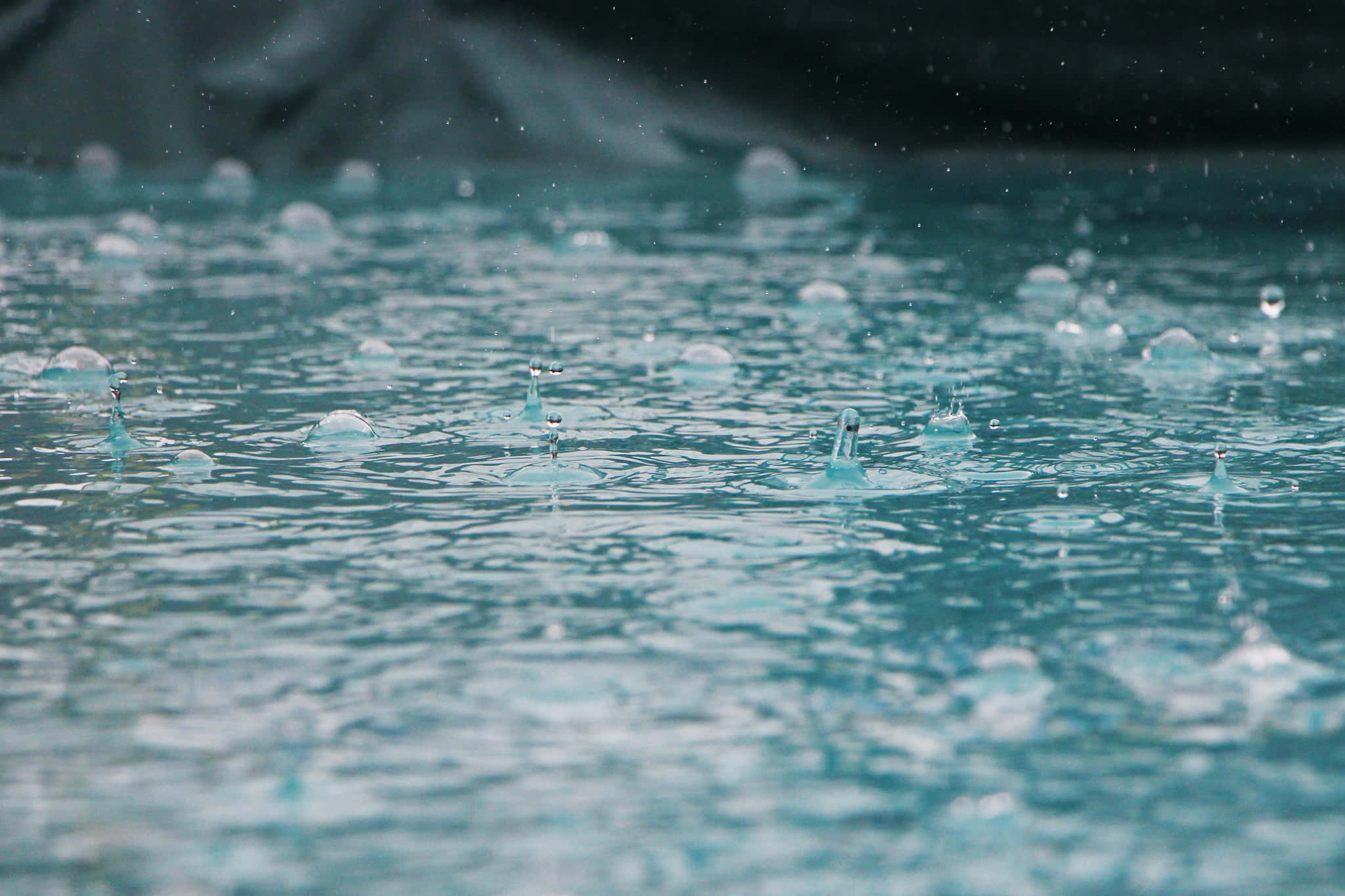 水 桌布 壁紙 — 雨滴在平靜的藍綠色水面上飛濺並泛起漣漪，背景有模糊的深色岩石。
