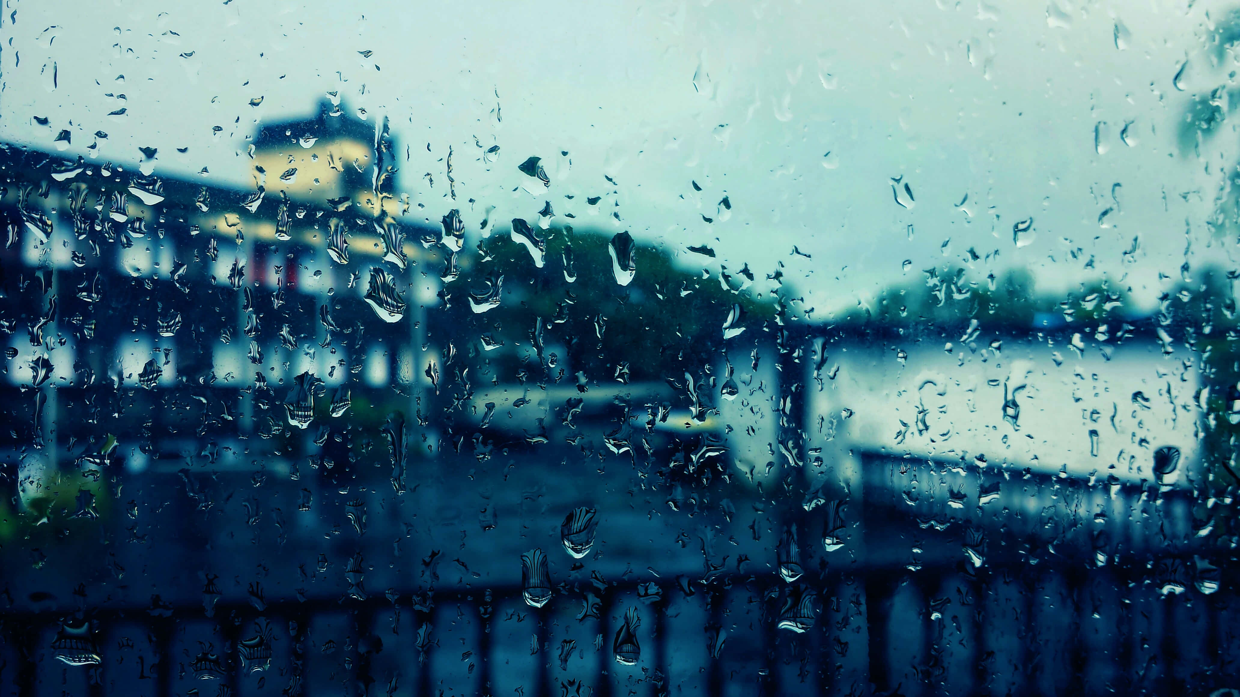 Building 桌布 壁紙 — 雨滴沿著玻璃窗流下，模糊了陰天下的柔和城市景觀。
