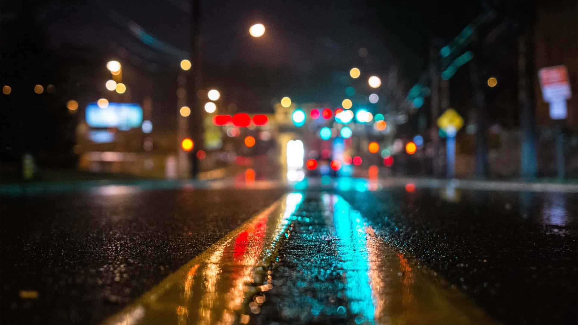  壁紙 — 雨濕的夜間城市街道，濕漉漉的瀝青和黃色車道標記上反射著發光的散景燈光。