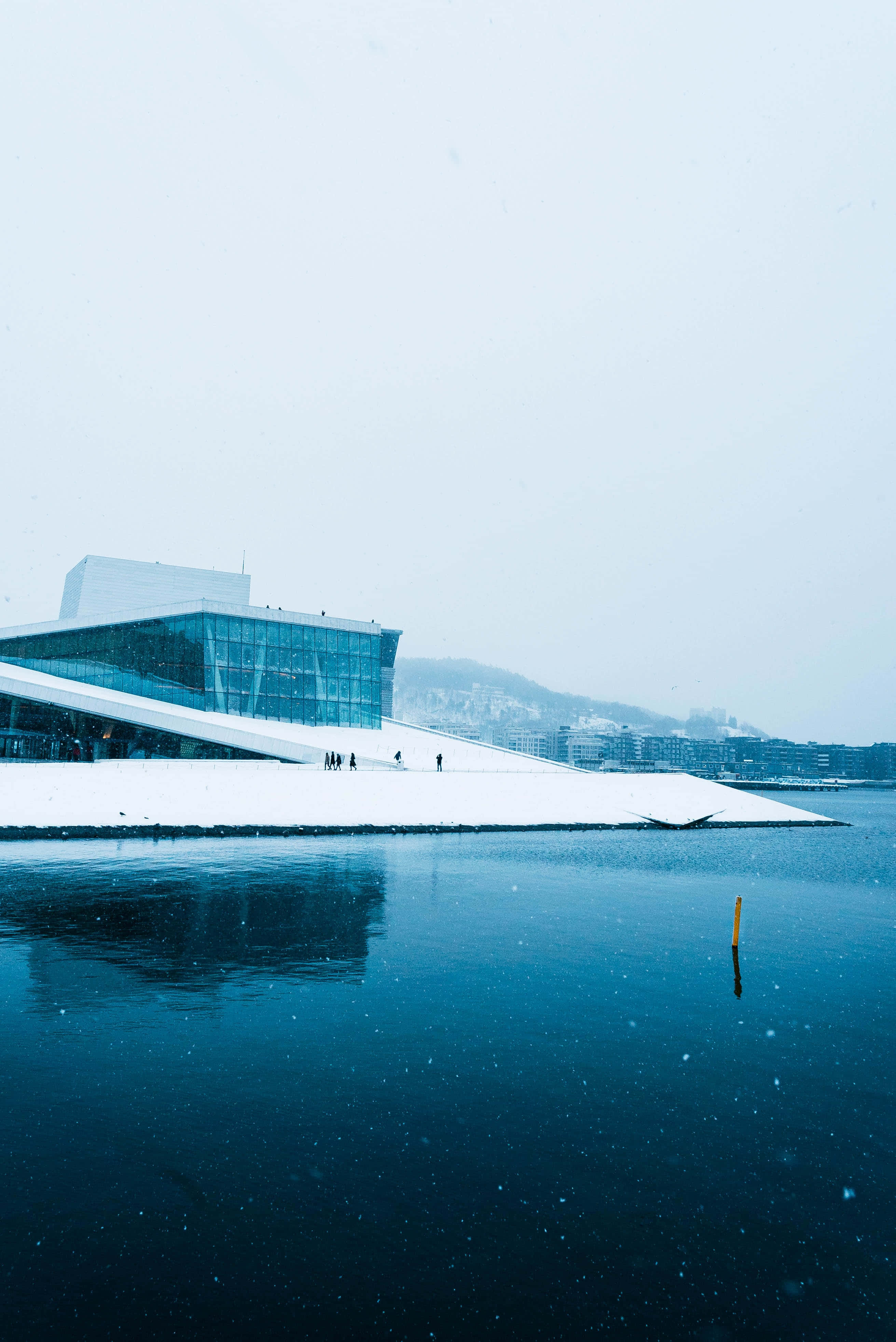 雪中奧斯陸歌劇院 壁紙 — 一座現代玻璃和積雪覆蓋的歌劇院在平靜的藍色水面上倒映，背景是白雪皚皚的冬日天空。