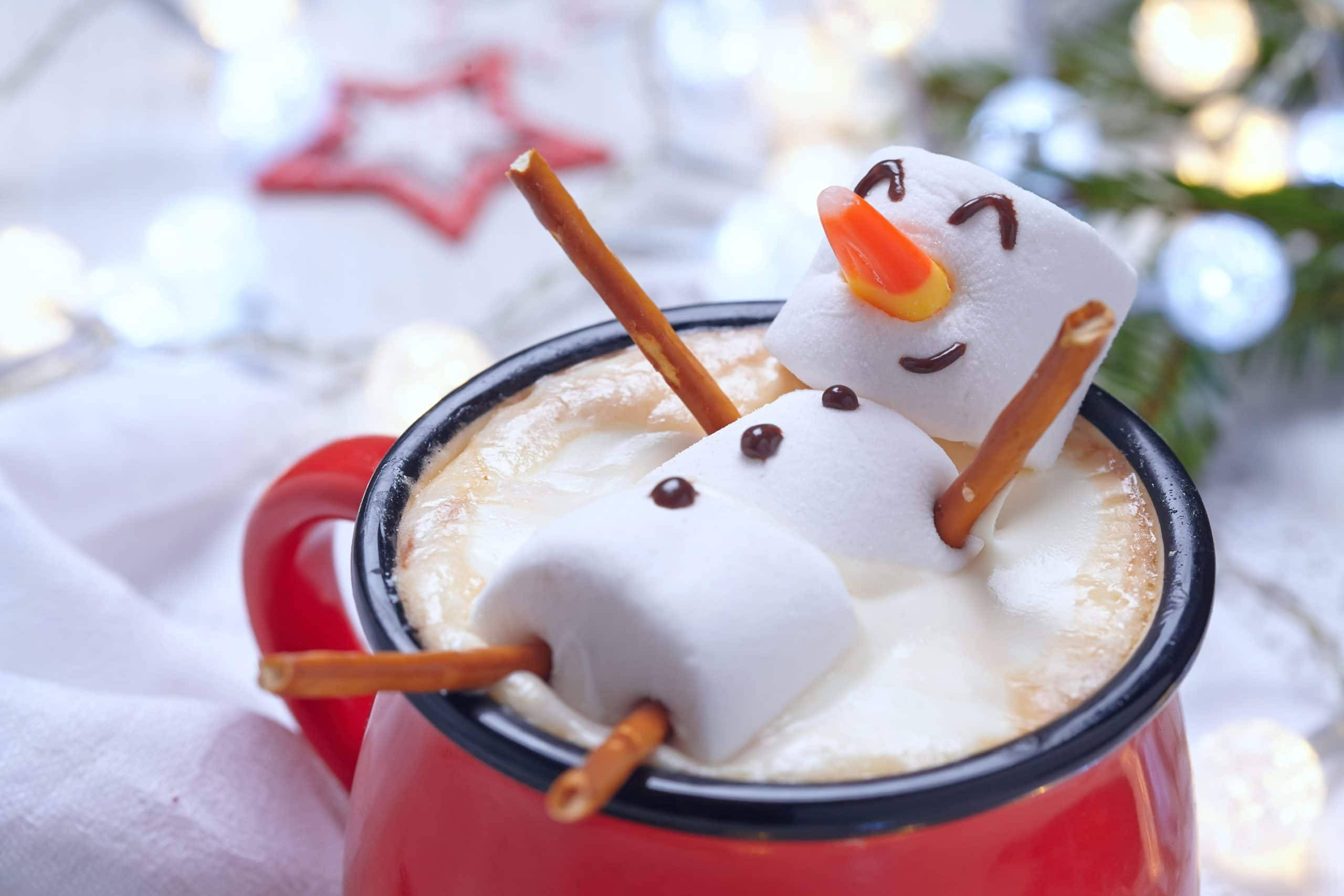雪人與暖飲 壁紙 — 一個舒適的紅色杯子裝著奶油熱朱古力，上面放著一個用椒鹽卷餅和糖果玉米製成的開心棉花糖雪人。