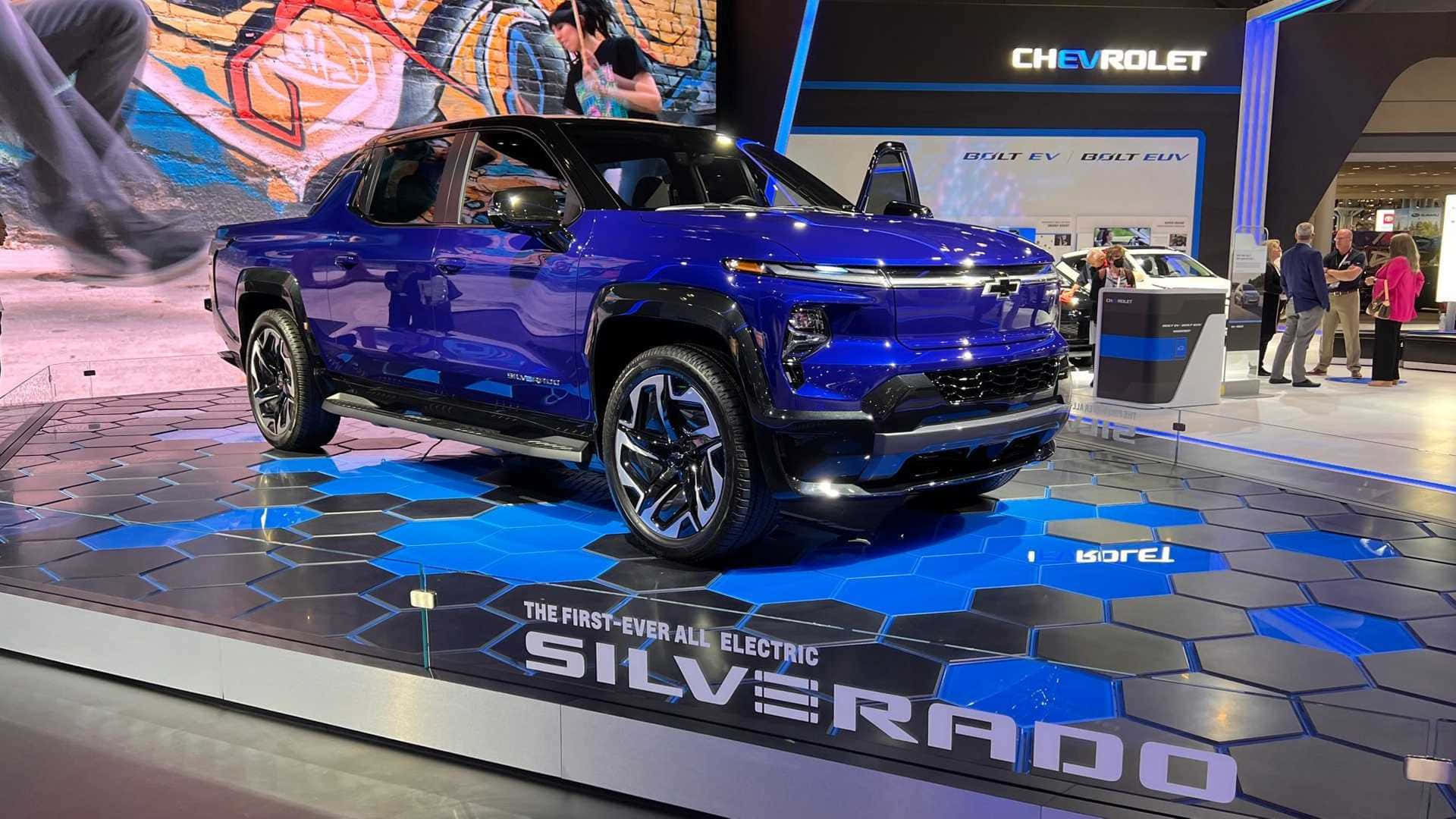 雪佛蘭經典與標誌性設計 壁紙 — 一輛閃亮的藍色雪佛蘭Silverado EV卡車，在六邊形藍黑舞台上展示，背景有人群。