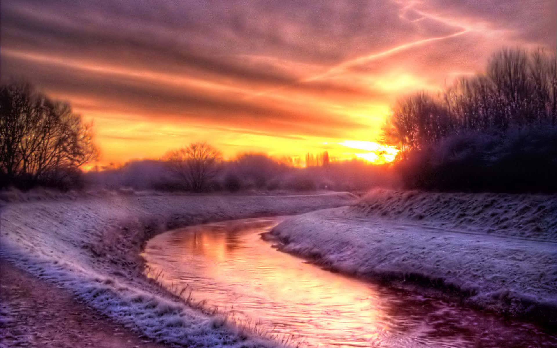 雪原上的冬季夕陽 壁紙 — 一條結冰的河流蜿蜒穿過雪景，在火紅的橙色和紫色日落天空下。