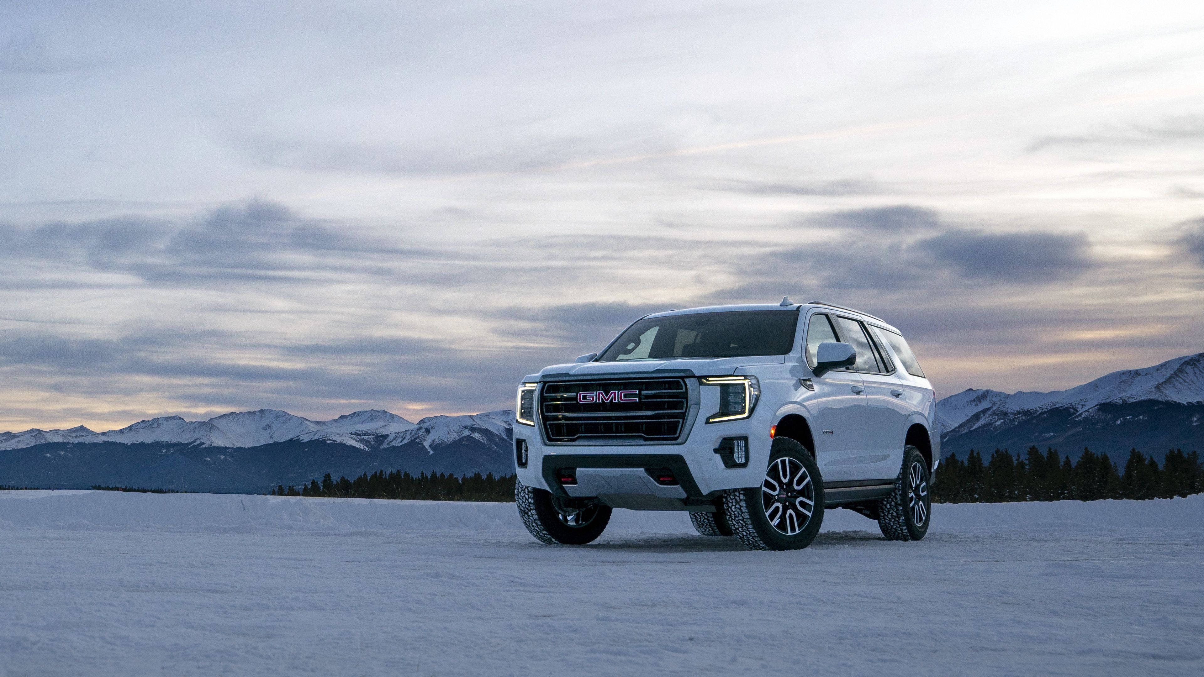 雪地中的GMC Yukon 壁紙 — 一輛白色GMC SUV矗立在廣闊的雪原上，背景是白雪覆蓋的山脈和多雲的黃昏天空。