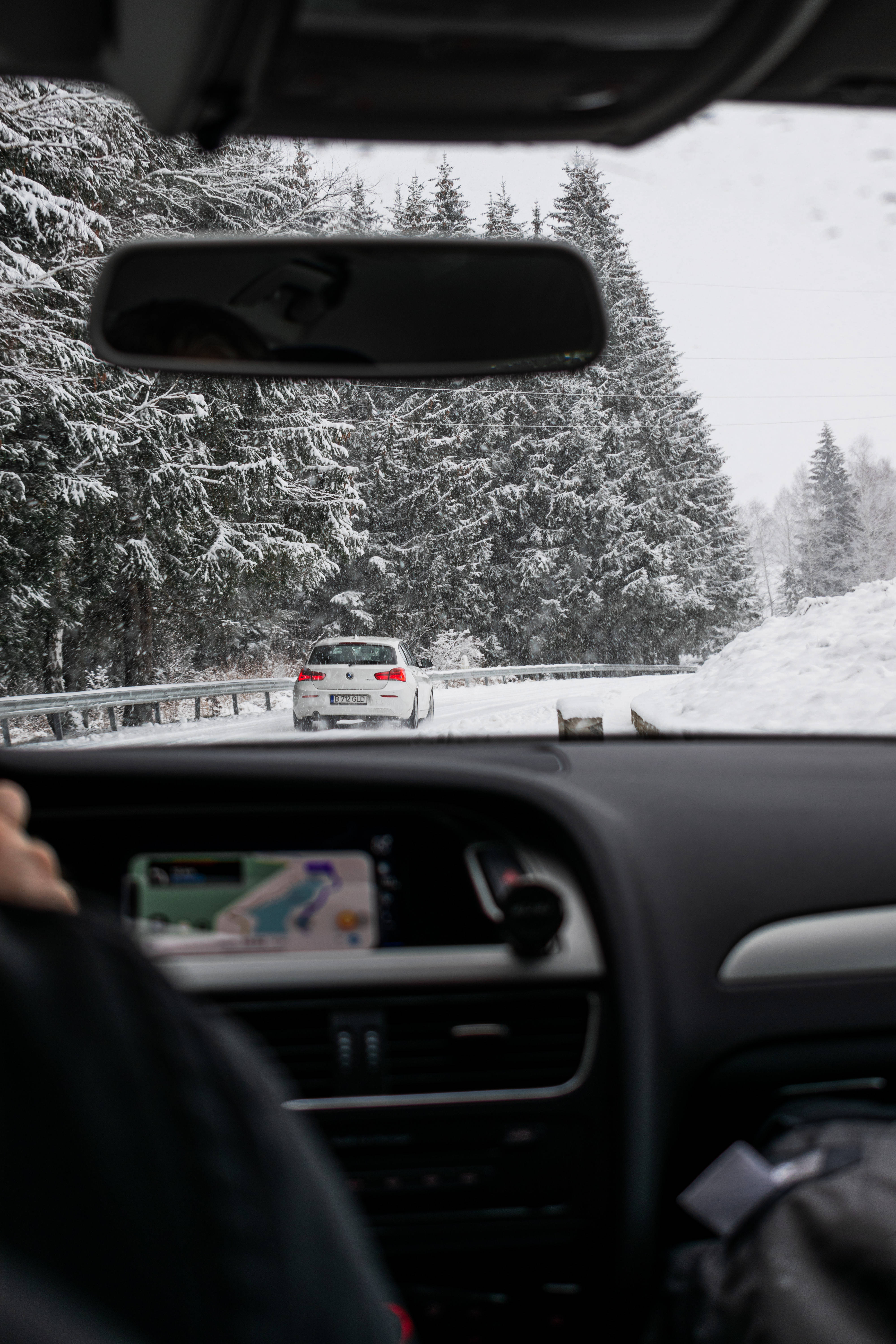 雪地公路驚豔手機壁紙 壁紙 — 從車內看到的雪地森林道路，前方有一輛白色汽車，道路兩旁是積雪的松樹。